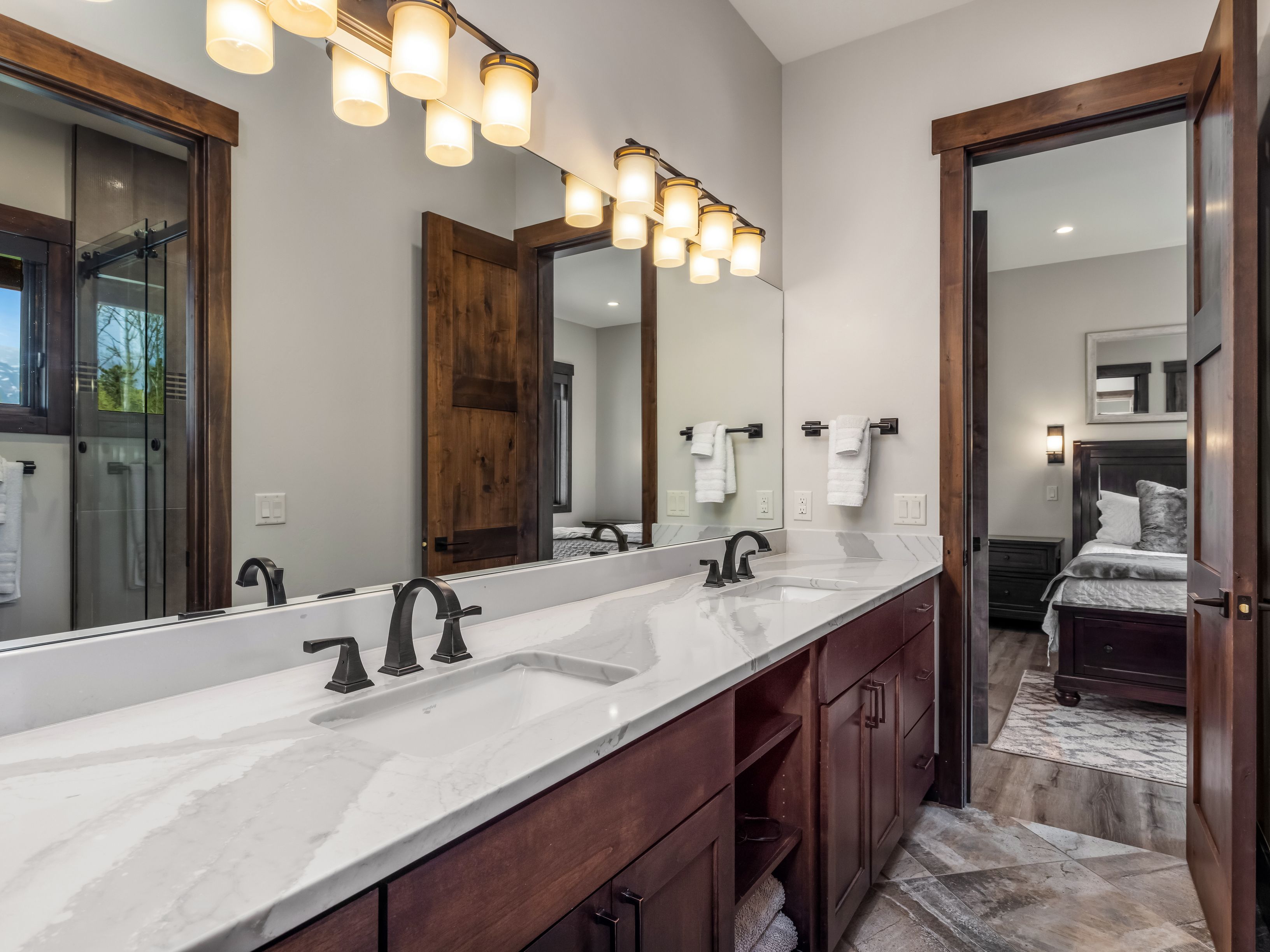 This primary bathroom features a dual-sink vanity with a light-colored stone countertop and dark wood cabinetry, complemented by dark metal fixtures. A large mirror spans the wall above the vanity, reflecting the adjacent bedroom and the warm lighting from the overhead fixtures. The space is finished with patterned tile flooring and rustic wood-trimmed doorways, creating a cohesive and sophisticated mountain-style aesthetic.