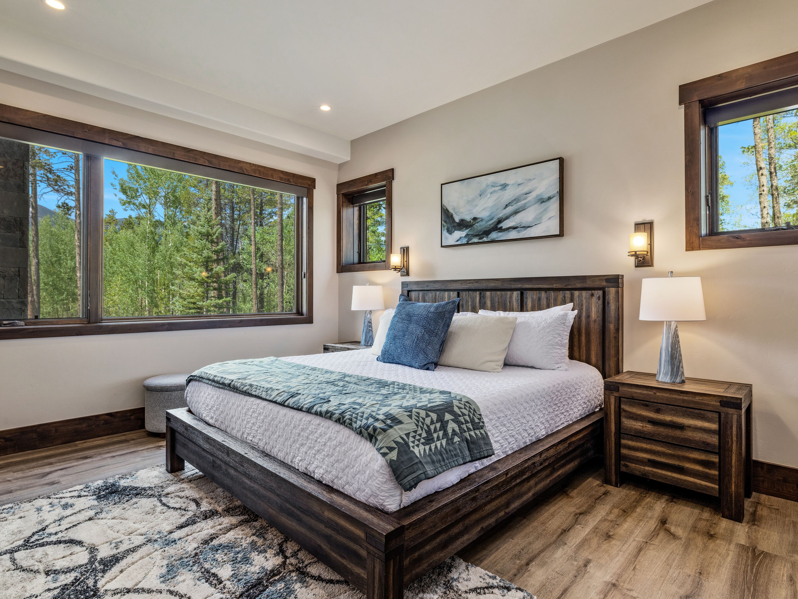 This primary bedroom features a rustic aesthetic with a dark wood bed frame and matching nightstands, complemented by light-toned walls and wood-look flooring. A large window provides an expansive view of the surrounding forest, while a patterned area rug and layered bedding add texture and comfort to the space. The room is illuminated by warm wall-mounted sconces and table lamps, creating a cozy and inviting atmosphere.