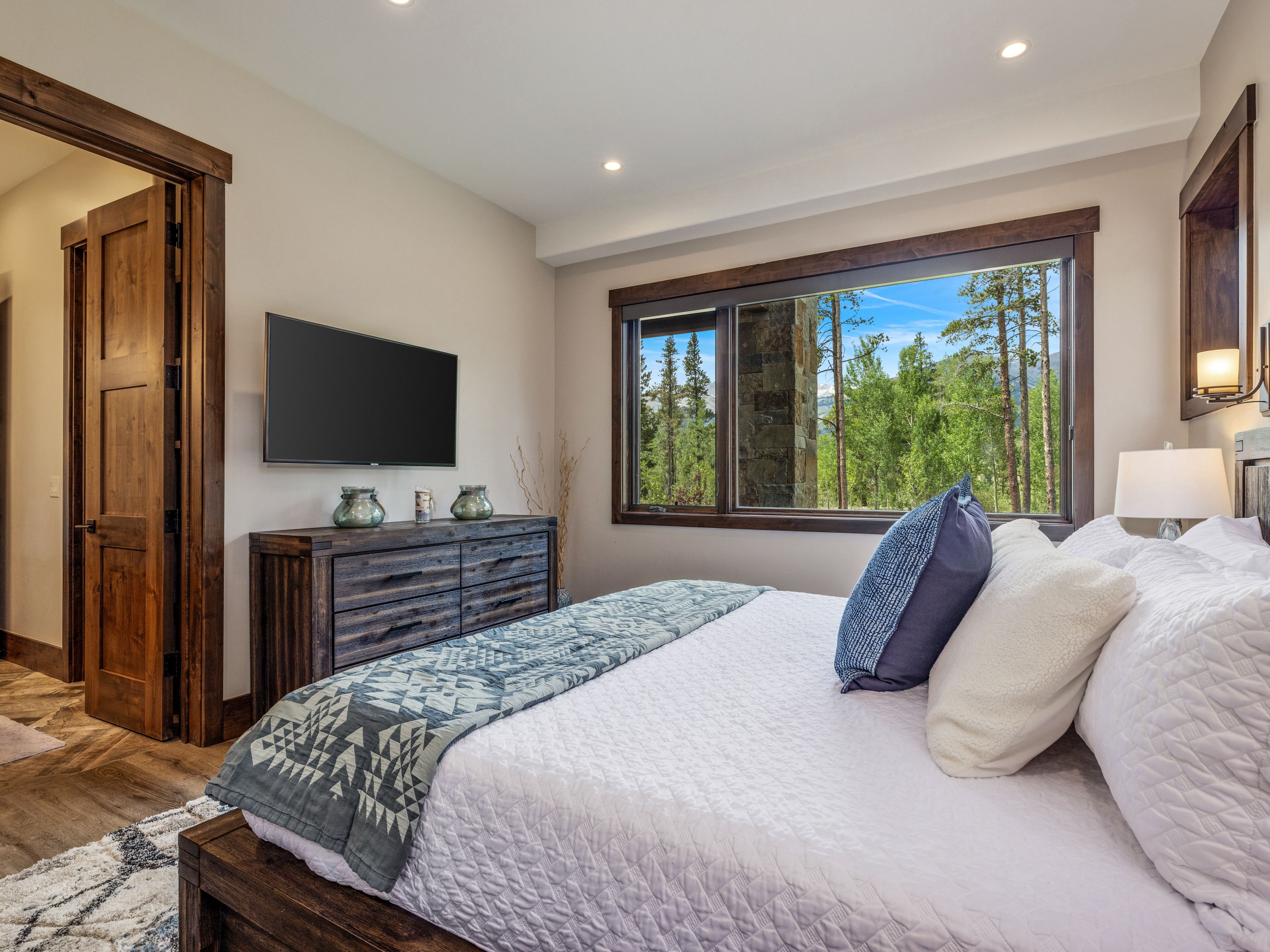 This cozy bedroom features a rustic aesthetic with dark wood furniture, including a sturdy bed frame and a matching dresser topped with decorative glass vases. A large window provides a scenic view of the surrounding forest and mountains, while a wall-mounted television offers entertainment. The room is accented with a patterned throw blanket and plush bedding, creating a warm and inviting atmosphere.