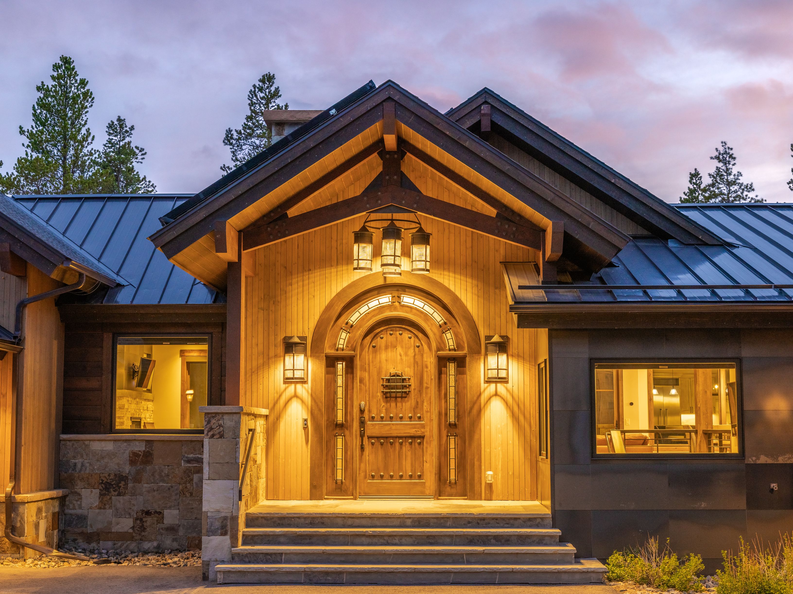 This striking mountain-style entryway features a grand arched wooden door framed by vertical wood siding and stone accents. The entrance is elevated by a set of stone steps and illuminated by warm, inviting exterior lanterns, creating a cozy and sophisticated atmosphere. The architectural design includes a prominent gabled roofline with exposed timber beams, set against a soft, twilight sky.