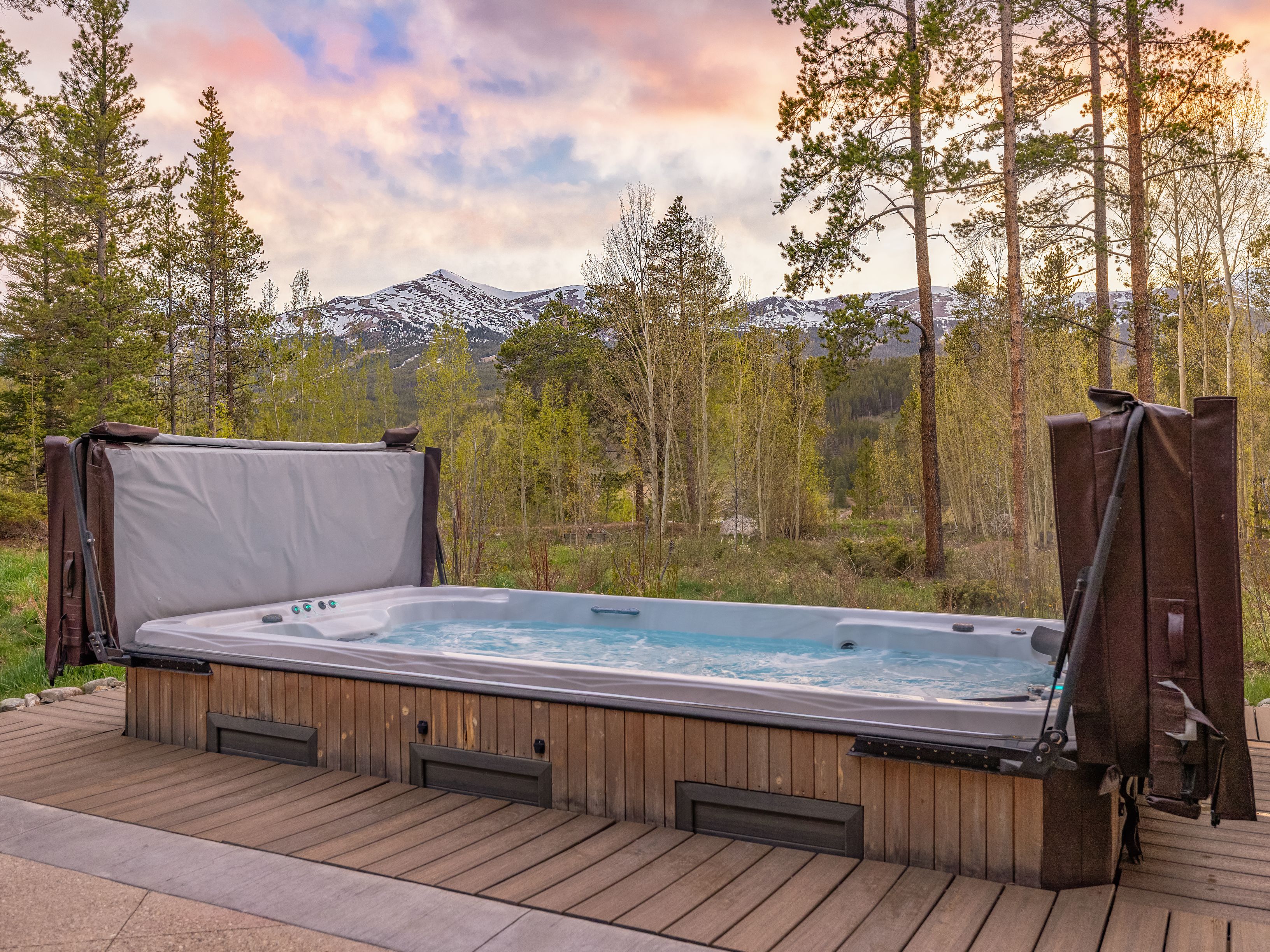 A large, rectangular swim spa is set into a wooden deck, featuring a light-colored interior and bubbling water. The spa is flanked by two upright, brown covers, and the surrounding landscape includes a lush green meadow, tall pine trees, and a majestic snow-capped mountain range in the distance. The scene is captured during a soft, colorful sunset, creating a serene and inviting atmosphere for outdoor relaxation.