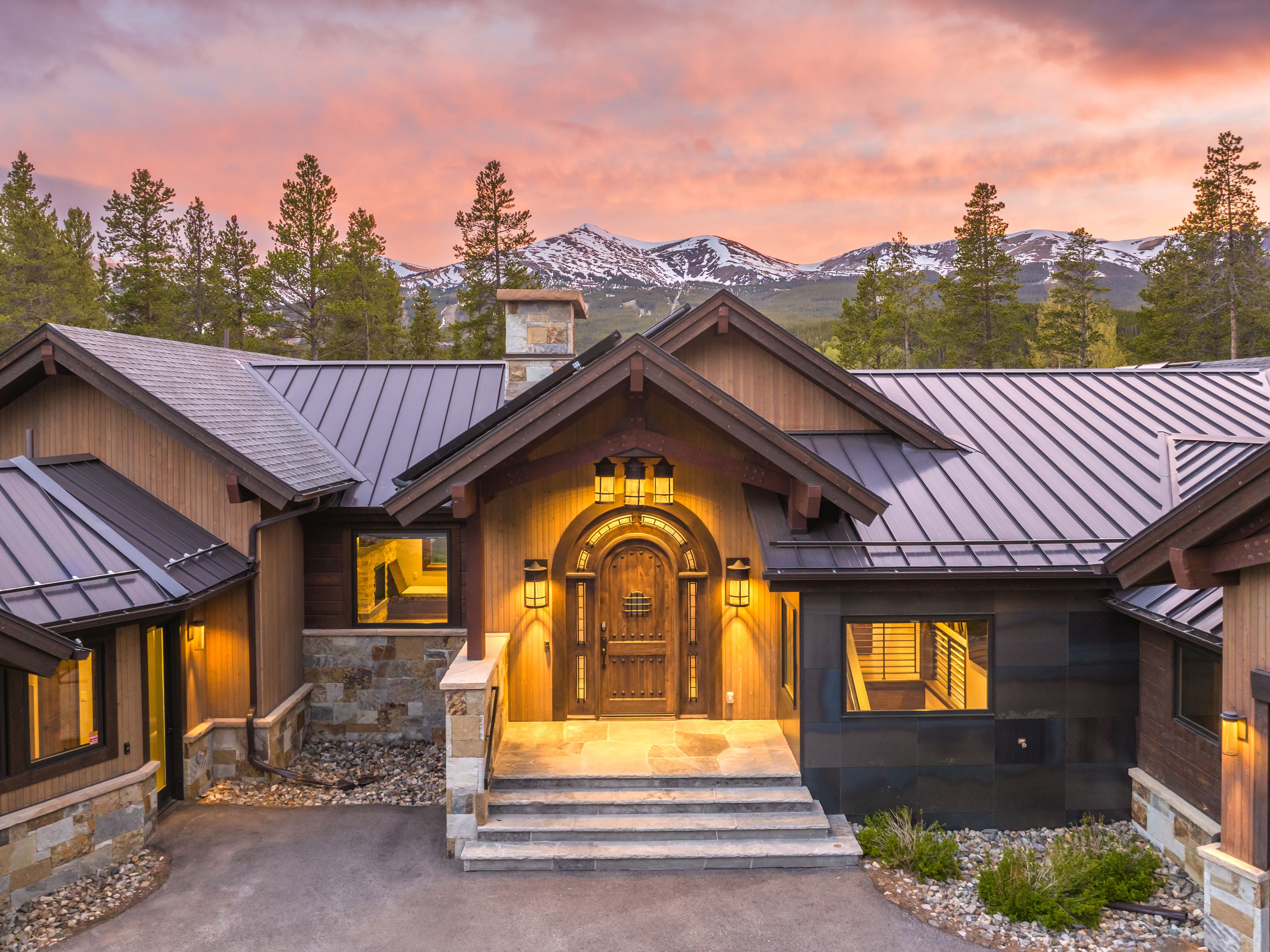 This stunning mountain-style home features a grand arched wooden front door framed by warm, inviting exterior lighting. The facade combines natural stone accents, vertical wood siding, and a dark metal roof, all set against a breathtaking backdrop of snow-capped peaks at sunset. The perspective is a slightly elevated eye-level shot, emphasizing the home's architectural symmetry and its harmonious integration with the surrounding alpine landscape.