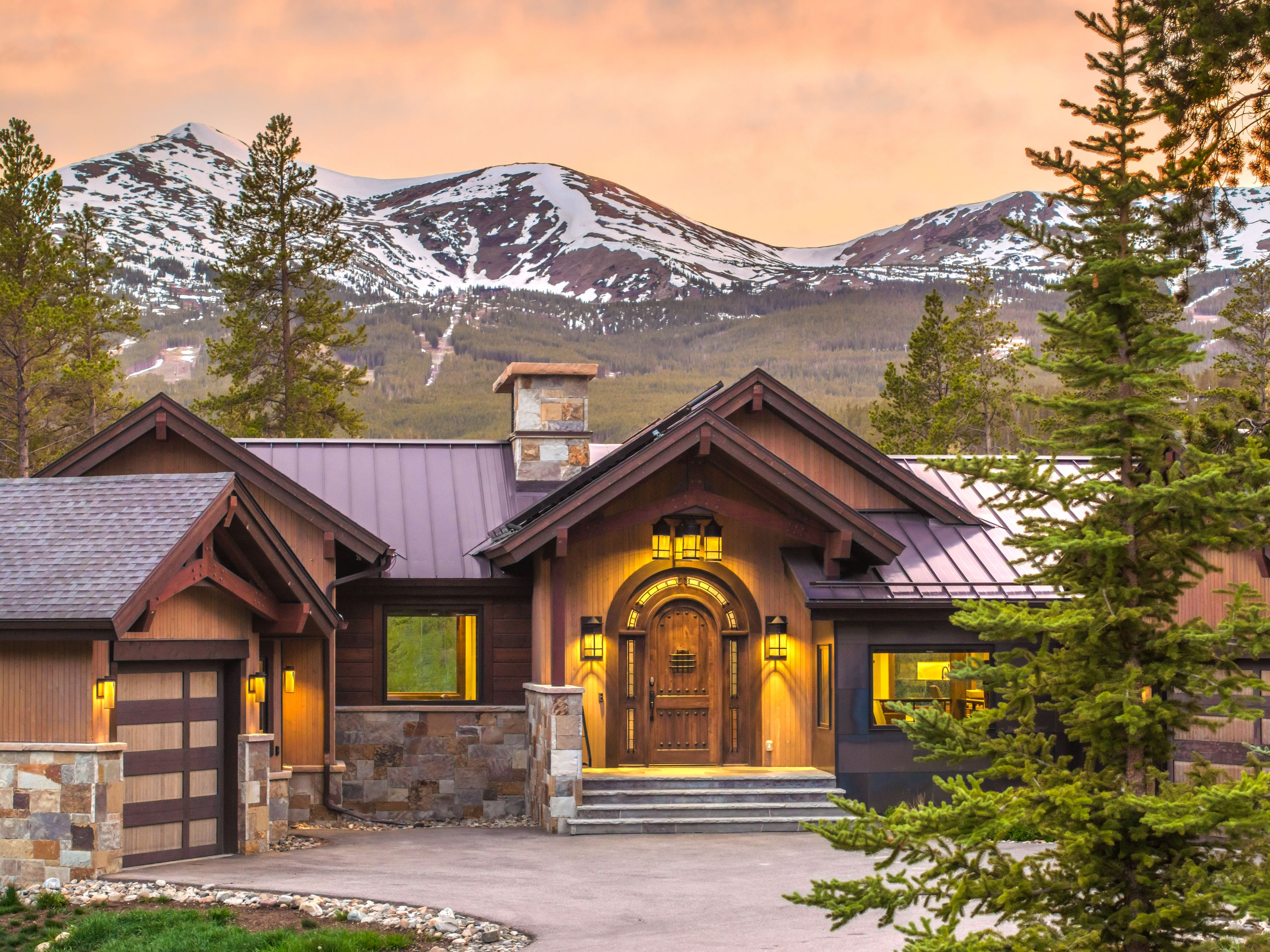 This mountain-style residence features a striking combination of stone masonry and warm-toned wood siding, complemented by a dark metal roof. The front entrance is highlighted by an arched wooden door and a stone-clad chimney rising above the roofline, all set against a dramatic backdrop of snow-capped peaks at sunset. The perspective captures the home's inviting curb appeal, framed by mature pine trees and a paved driveway.