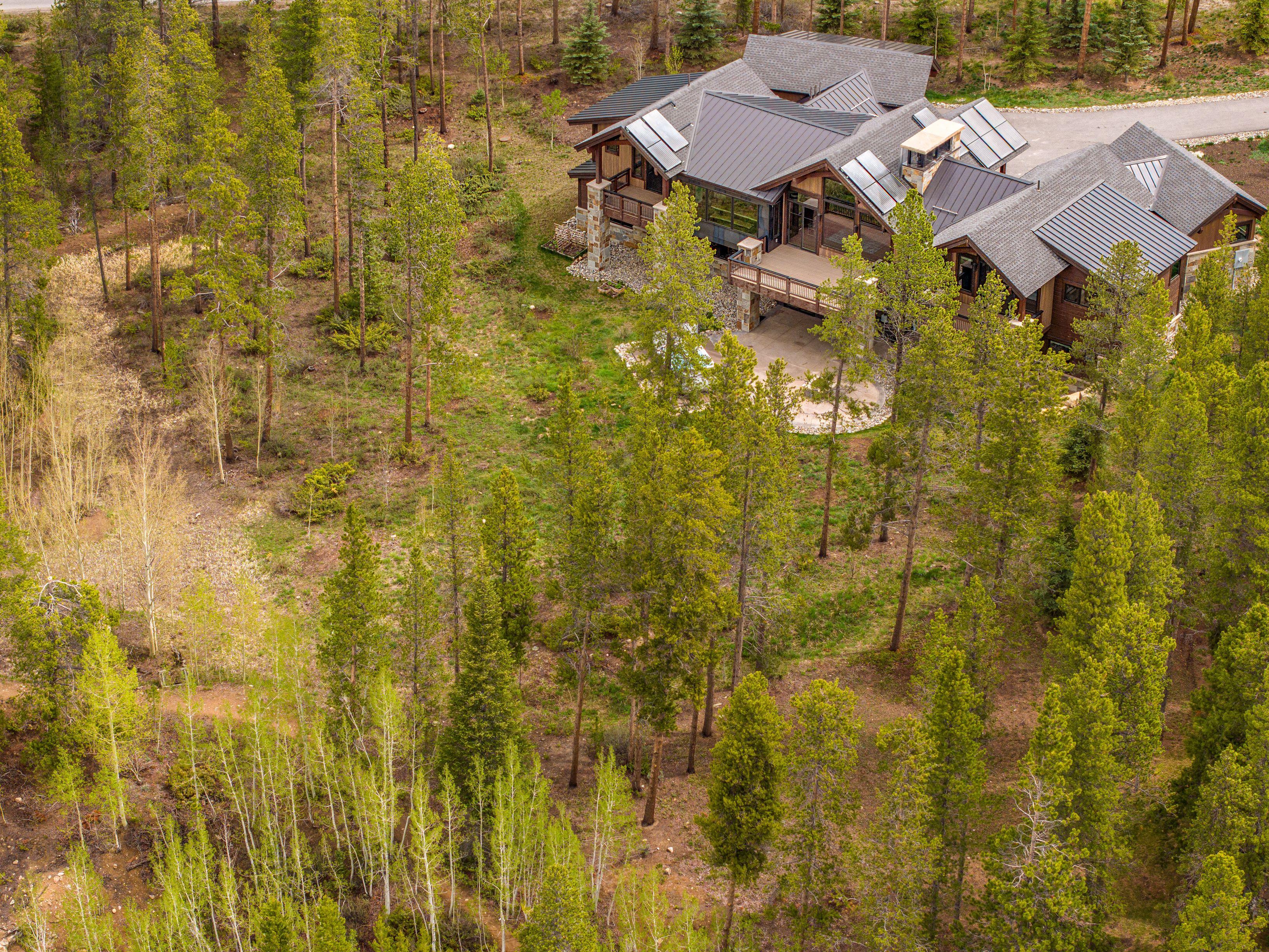This high-angle aerial view captures a sprawling, multi-gabled mountain home nestled within a dense forest of pine and aspen trees. The residence features a dark metal roof, stone and wood siding, and a spacious elevated deck overlooking a stone-paved patio area below. The surrounding landscape is natural and wooded, emphasizing a secluded, rustic retreat aesthetic.