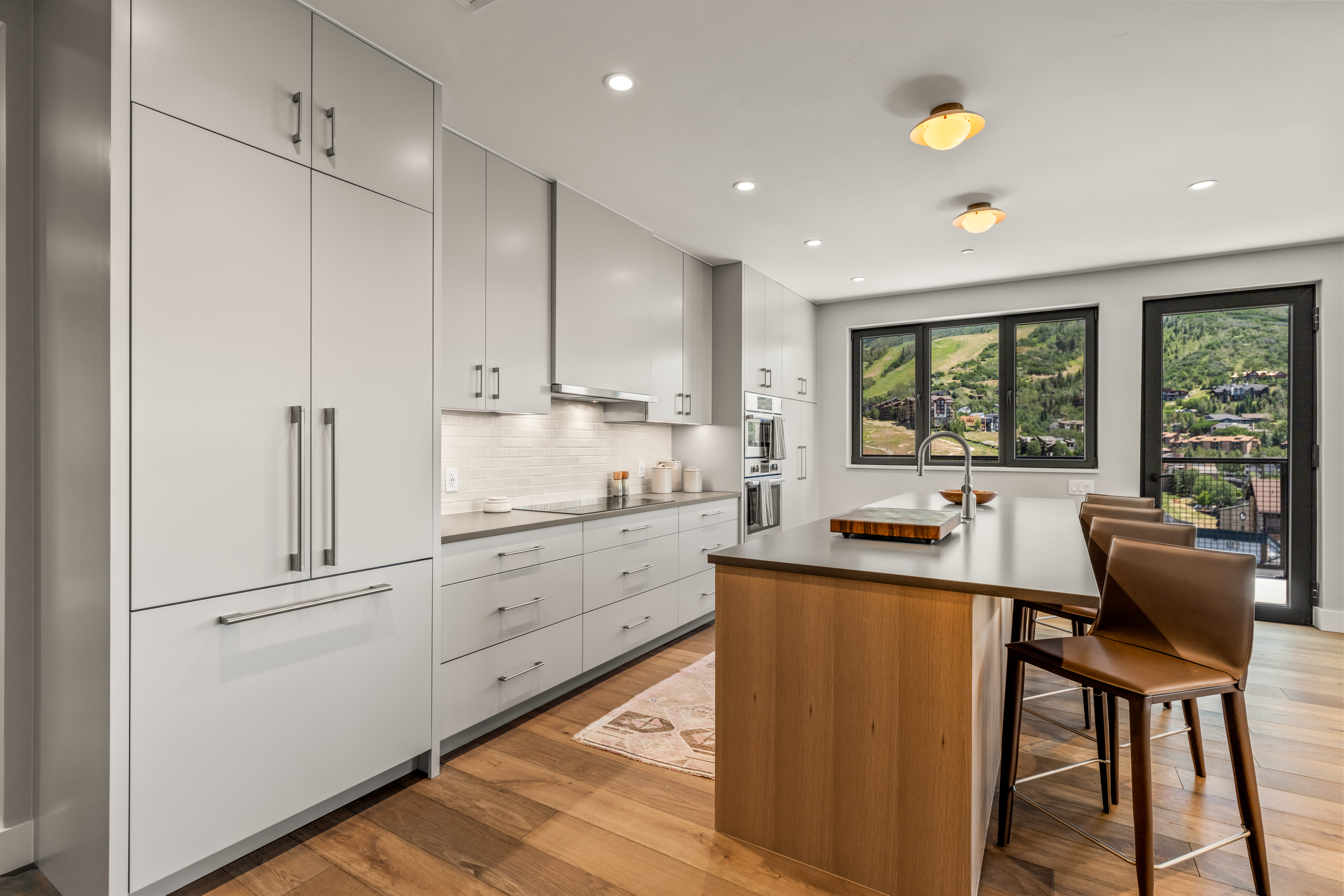 This modern kitchen features sleek, light-colored cabinetry paired with a central island that has a warm wood base and a dark countertop. The space is illuminated by recessed lighting and two distinctive circular ceiling fixtures, while a large window provides a scenic view of a mountain landscape. A patterned runner rug adds texture to the wood flooring, creating a clean and inviting atmosphere.
