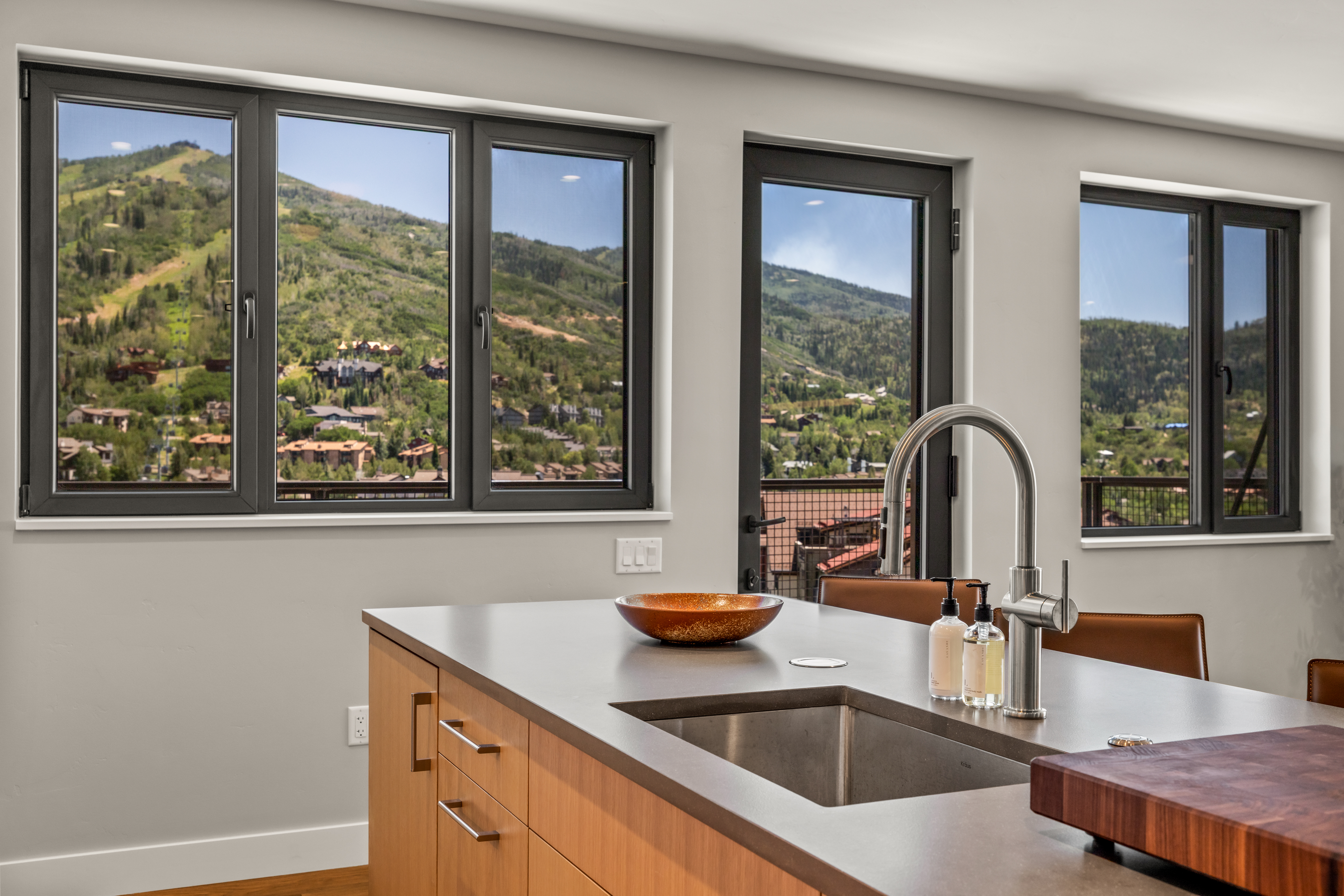 This modern kitchen features a central island with a stainless steel sink, a sleek faucet, and a dark stone countertop. The cabinetry is crafted from light wood with clean lines, and the space is framed by large windows that offer a panoramic view of a mountain landscape. The perspective is eye-level, highlighting the functional workspace and the seamless connection between the interior and the scenic outdoor environment.