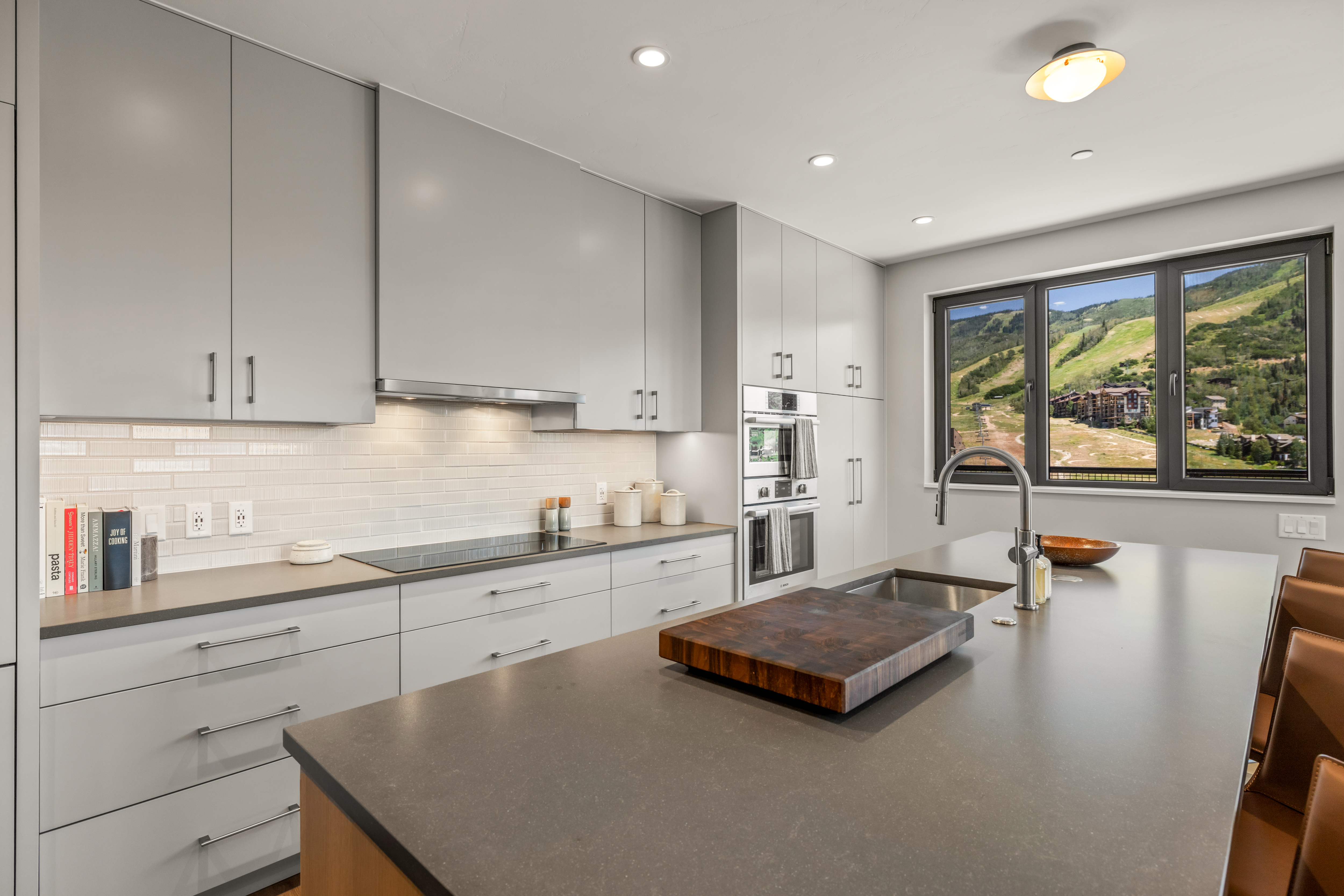 This modern kitchen features sleek, light gray cabinetry paired with dark stone countertops and a matching large central island. A built-in wall oven and cooktop are integrated into the cabinetry, while a large window above the sink provides a scenic view of a mountain landscape. The space is illuminated by recessed lighting and a simple ceiling fixture, creating a clean, contemporary aesthetic.