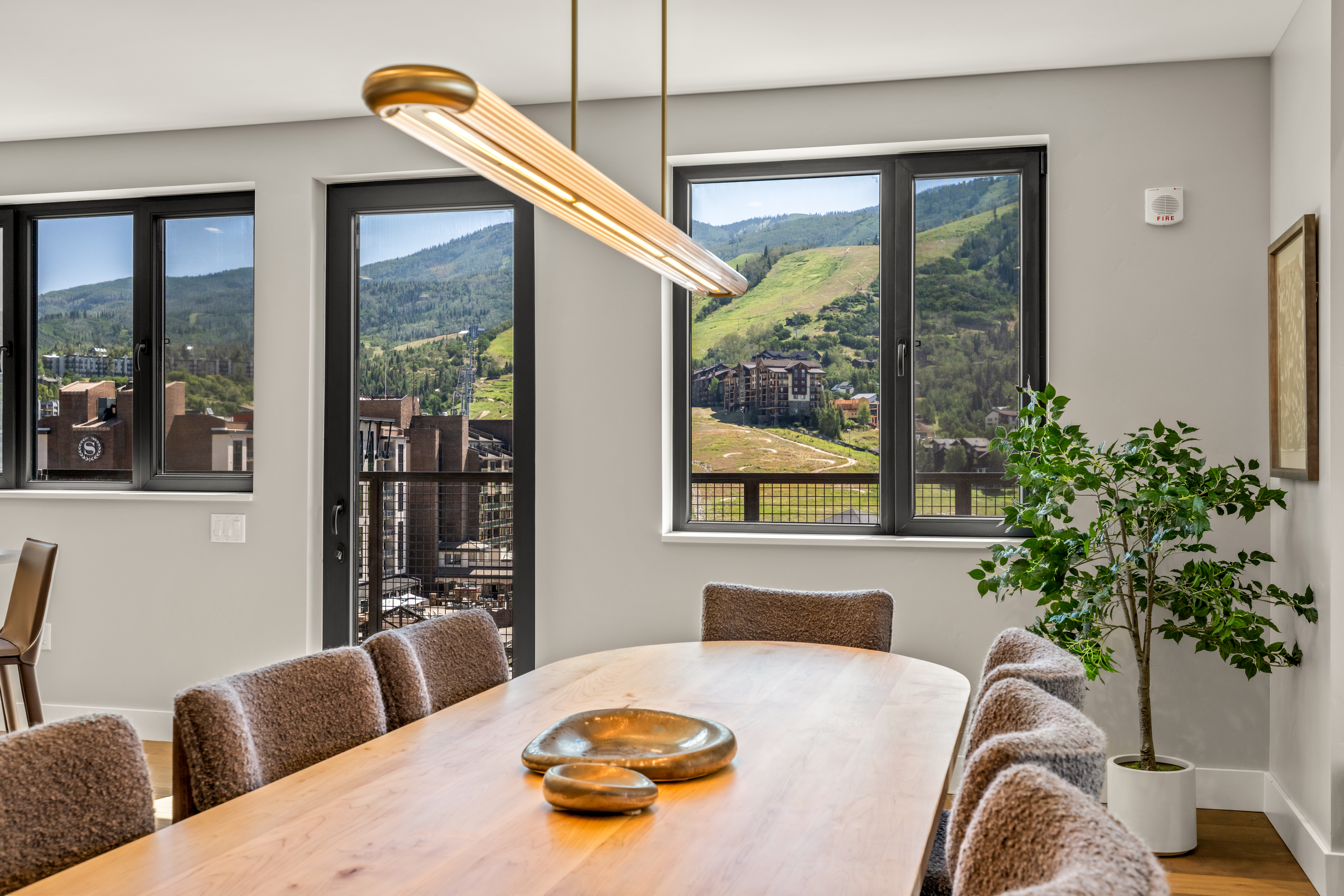 This bright dining room features a long, light wood table surrounded by textured, upholstered chairs, centered beneath a modern, linear gold-toned light fixture. Large windows and a glass door provide expansive views of a mountain landscape and ski resort area. A potted plant adds a touch of greenery to the corner, creating a serene and inviting atmosphere.