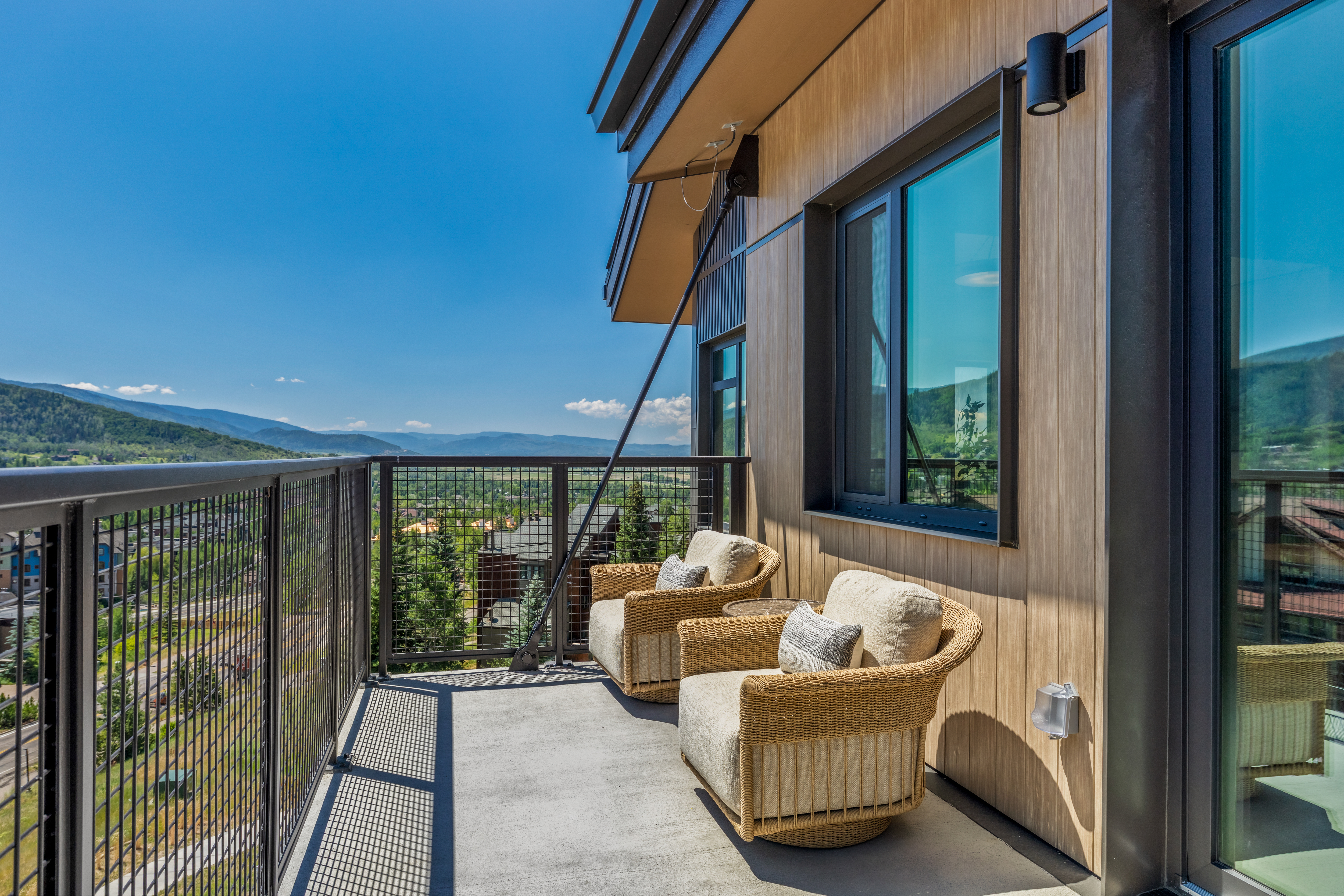 This modern balcony features two comfortable wicker armchairs with neutral cushions, providing a perfect spot to enjoy the expansive mountain views. The space is finished with a sleek metal railing and light wood-toned siding, creating a contemporary and inviting outdoor living area. The perspective captures the open-air setting, highlighting the seamless transition between the interior living space and the scenic landscape.