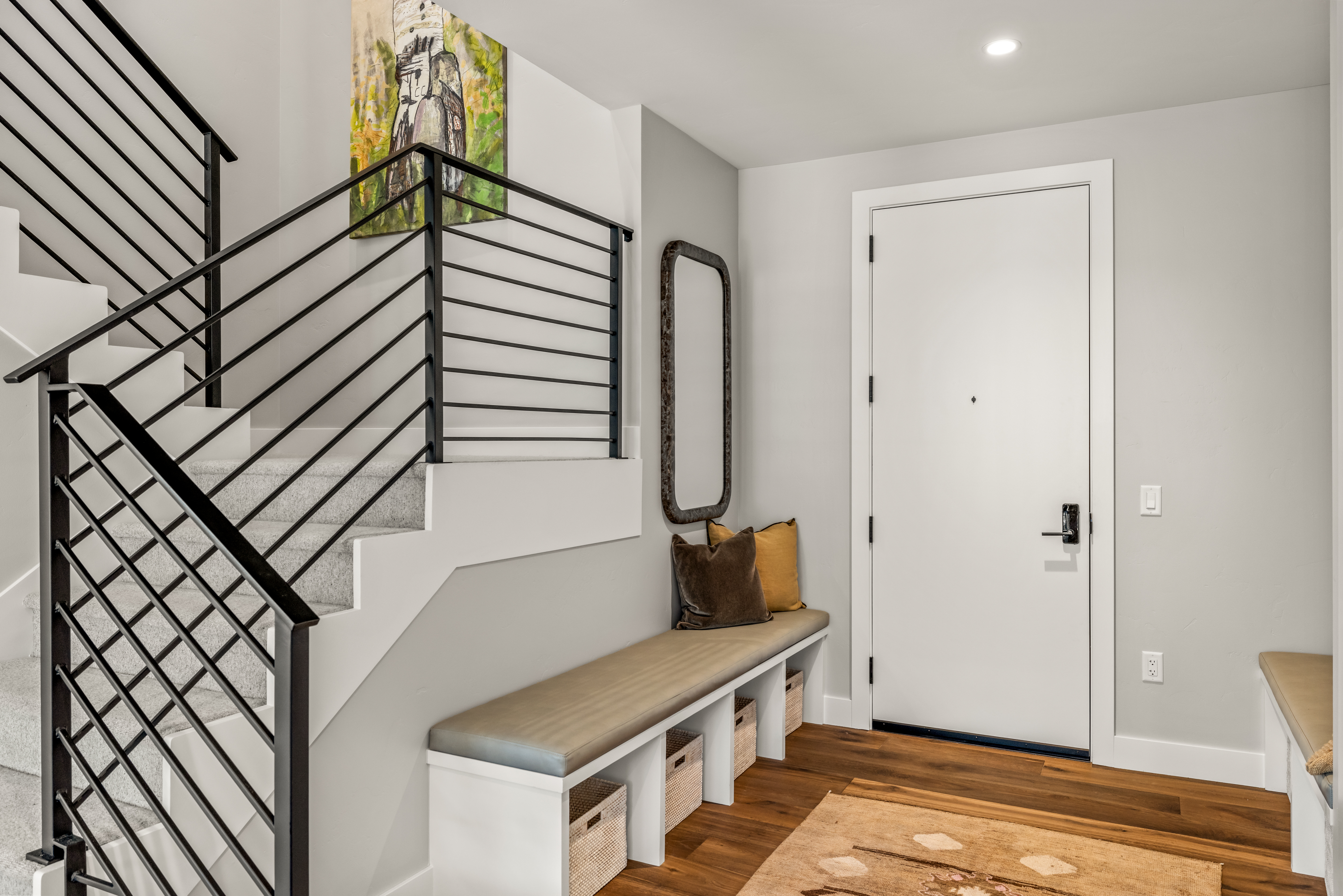 This bright entryway features a modern staircase with black metal railings and light-colored carpeted steps. A built-in bench with integrated storage baskets sits against the wall, topped with decorative pillows and a tall, narrow mirror. The space is finished with light gray walls, warm wood flooring, and a white front door, creating a clean and welcoming transition area.