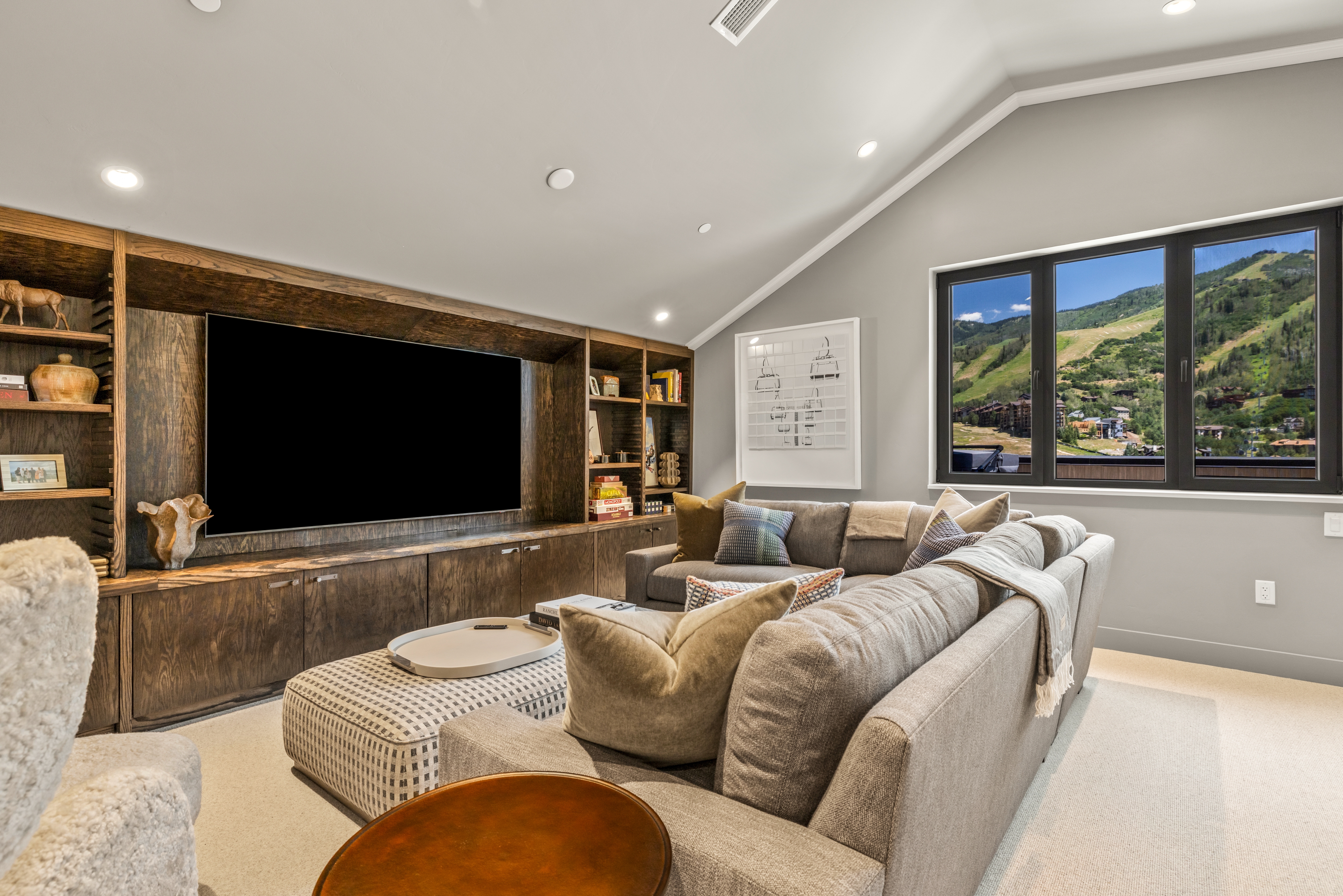 This cozy living room features a large, dark wood media console with integrated shelving that houses a television and decorative items. A plush, L-shaped sectional sofa with various throw pillows faces the media wall, complemented by a patterned ottoman and a small round side table in the foreground. Large windows on the right provide a scenic view of a mountain landscape, while the vaulted ceiling and neutral color palette create an inviting and spacious atmosphere.
