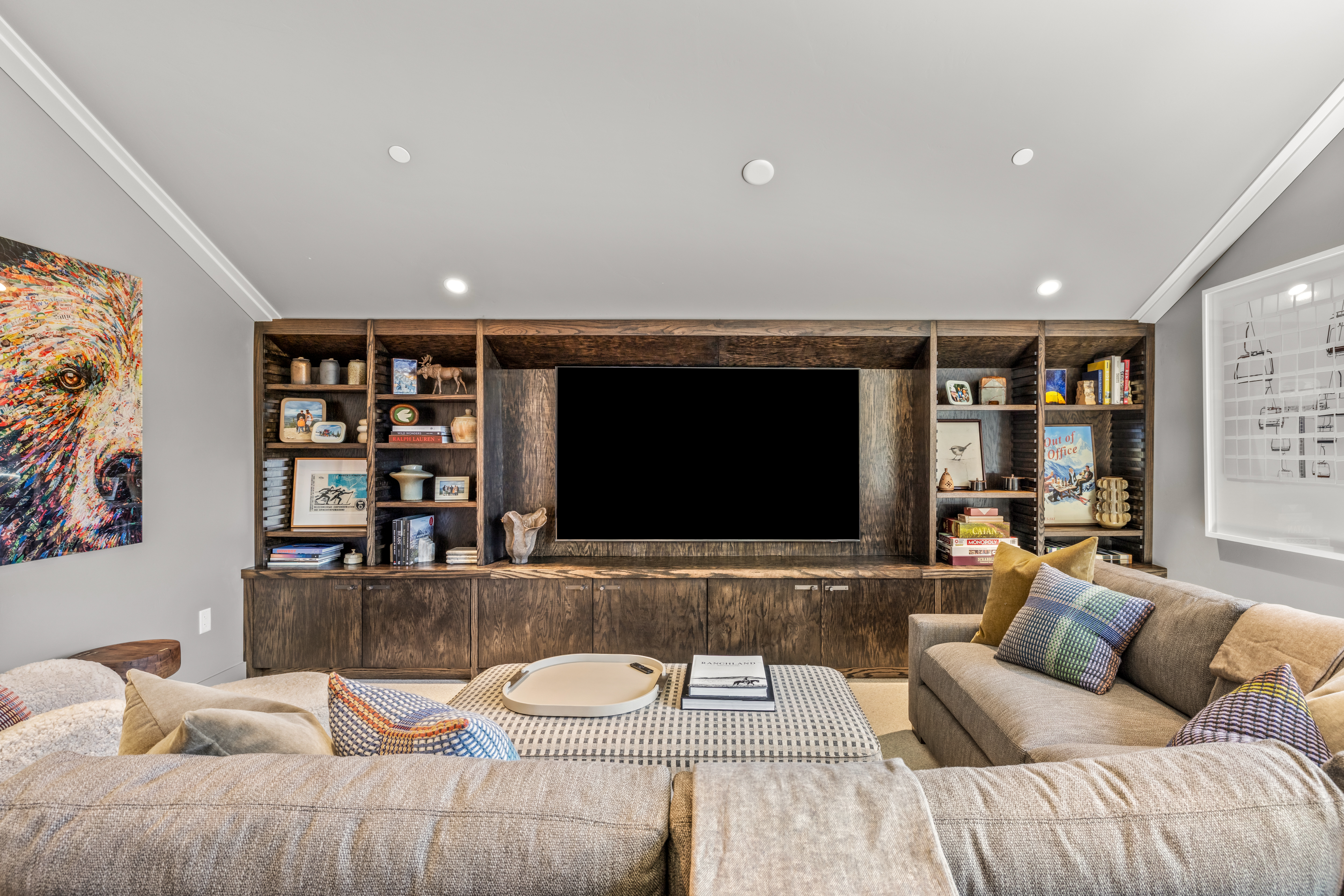 This inviting living room features a large, dark wood built-in media center that serves as the focal point, housing a television and various decorative items on open shelving. A spacious, neutral-toned sectional sofa with patterned accent pillows and a matching ottoman creates a comfortable seating area. The room is accented by a vibrant, colorful bear portrait on the left wall and soft recessed lighting, contributing to a warm and cozy atmosphere.