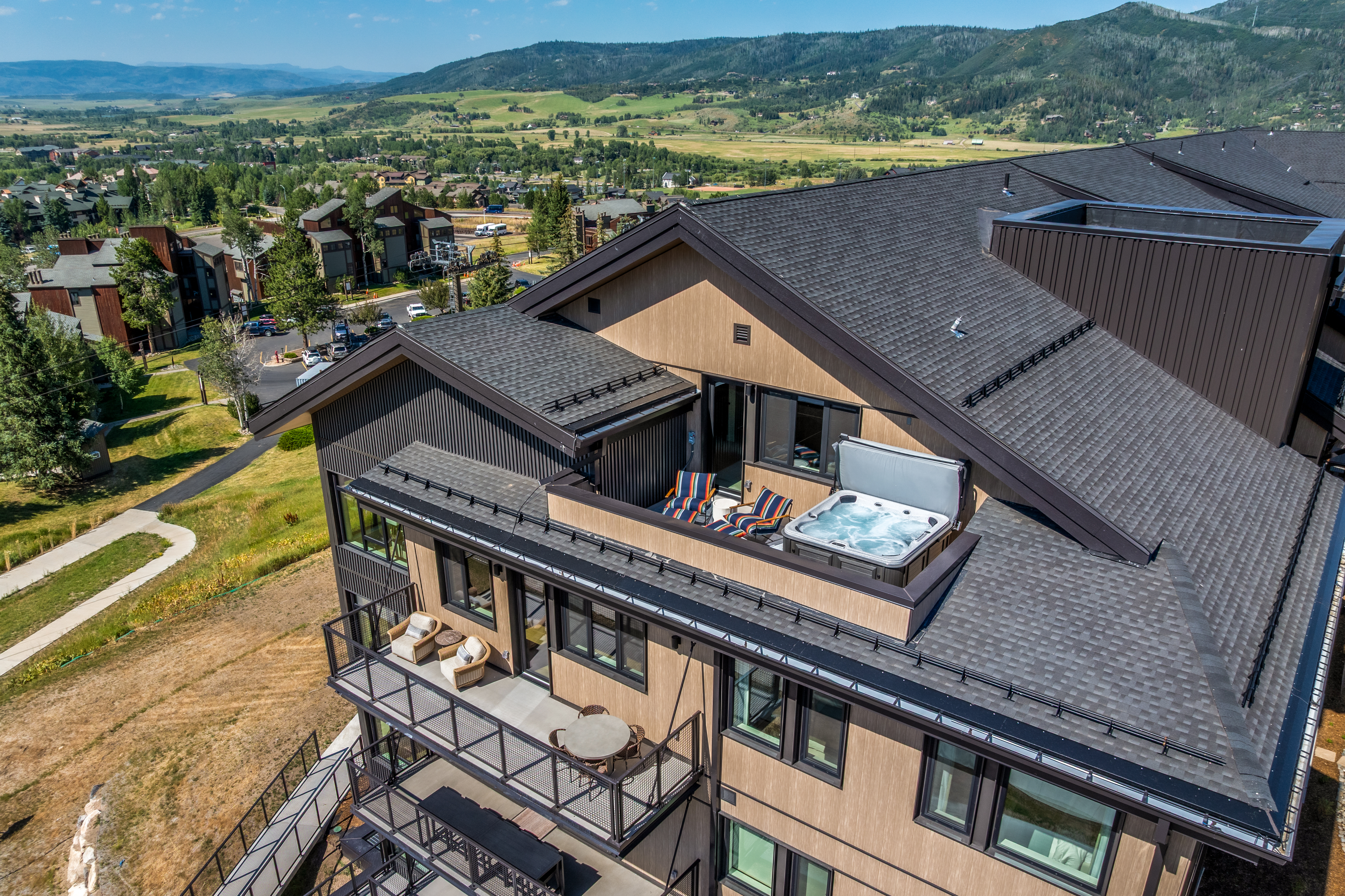 This high-angle aerial shot captures a modern multi-story residential building featuring a prominent rooftop terrace equipped with a hot tub and lounge seating. Below, a spacious balcony offers additional outdoor living space with a dining set and comfortable chairs, all overlooking a scenic valley landscape. The architecture showcases a contemporary mountain aesthetic with dark roofing, wood-toned siding, and clean, geometric lines.