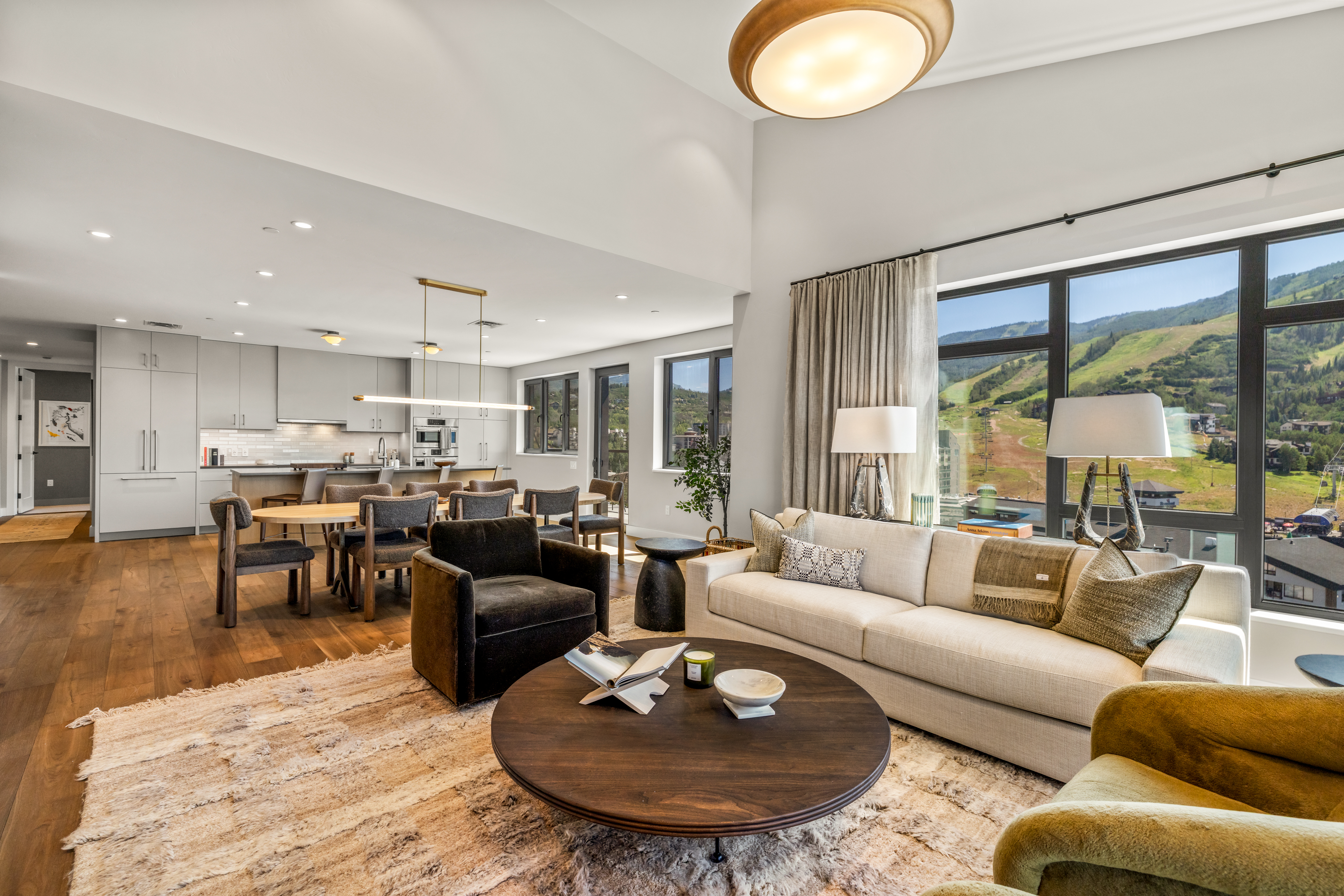 This open-concept living and dining area features a sophisticated, modern aesthetic with warm wood flooring and a neutral color palette. A large, circular wooden coffee table anchors the seating area, which includes a plush sofa and a dark armchair, all set against a backdrop of expansive windows showcasing mountain views. The adjacent dining space is illuminated by a sleek, linear pendant light, creating a seamless flow between the kitchen and the main living area.