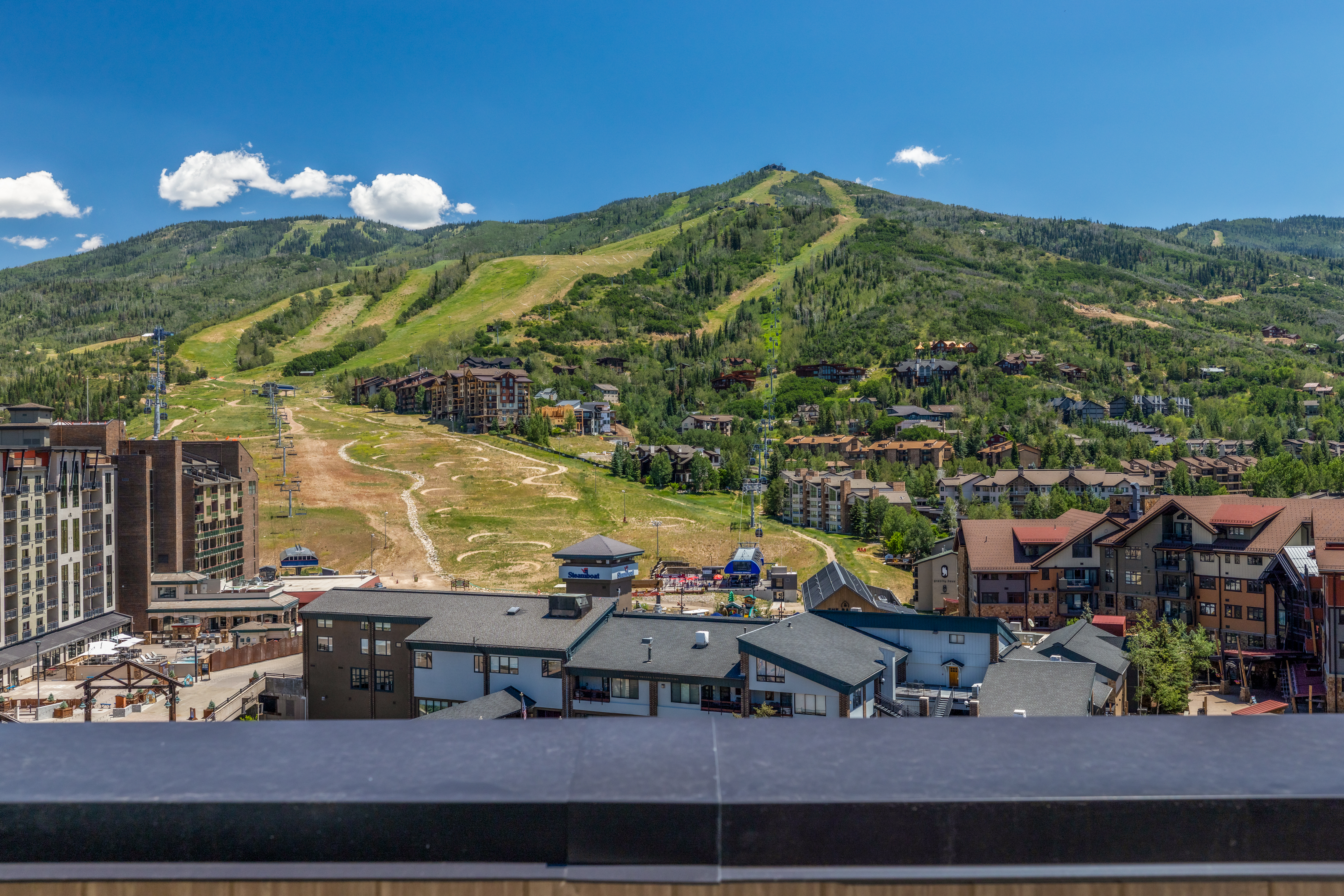 This elevated perspective captures a sweeping panoramic view of a mountain resort from a balcony. The foreground features a dark, horizontal railing, while the mid-ground reveals a complex of multi-story residential buildings and a ski base area. In the background, a lush, green mountain slope with ski runs stretches toward a clear blue sky, creating an inviting and scenic atmosphere.