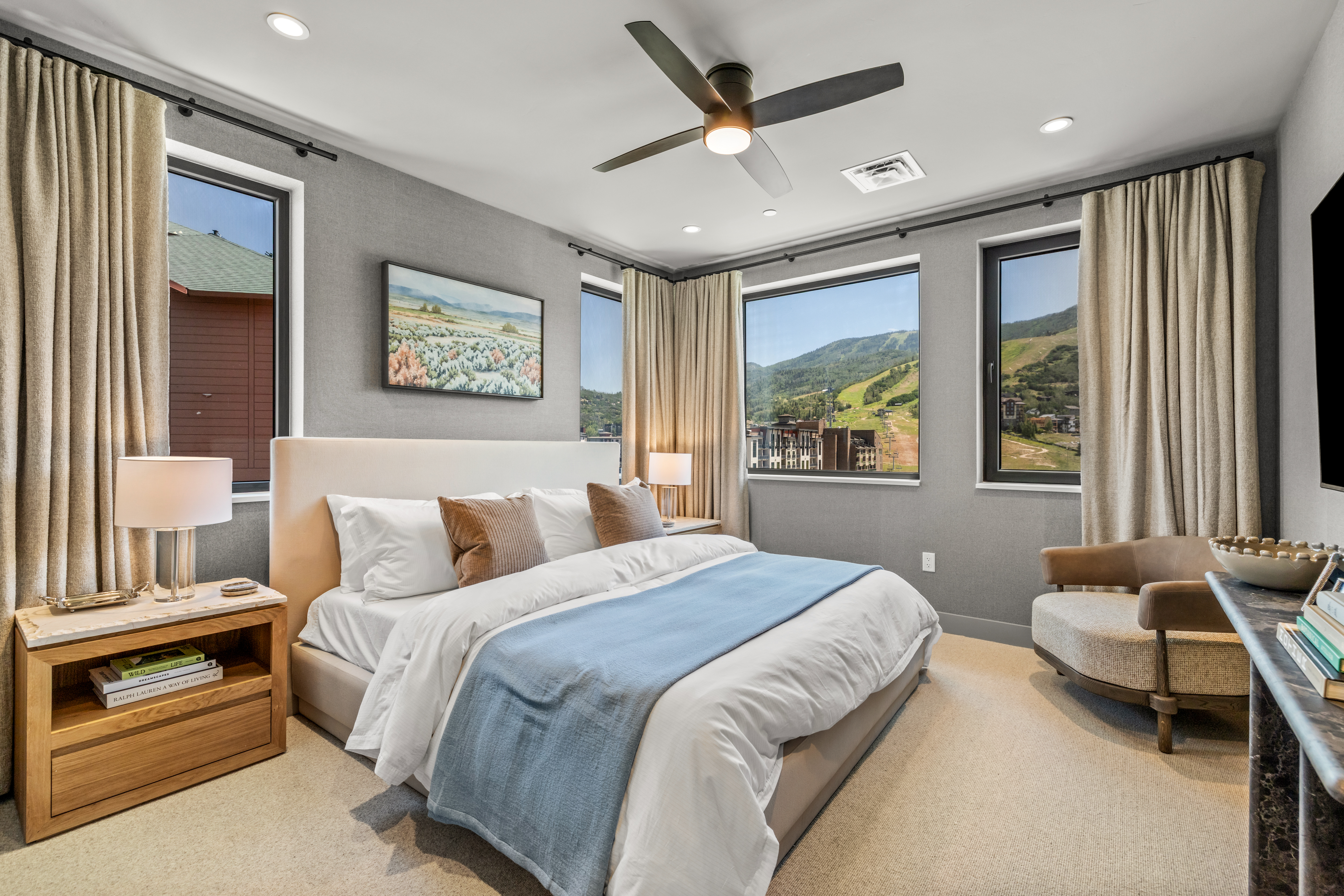 This primary bedroom features a neutral color palette with textured gray walls and a plush upholstered headboard. The room is anchored by a bed dressed in white linens with a soft blue throw, flanked by light wood nightstands and matching lamps. Large windows provide expansive views of the surrounding mountain landscape, while a comfortable armchair sits in the corner, creating a serene and inviting retreat.