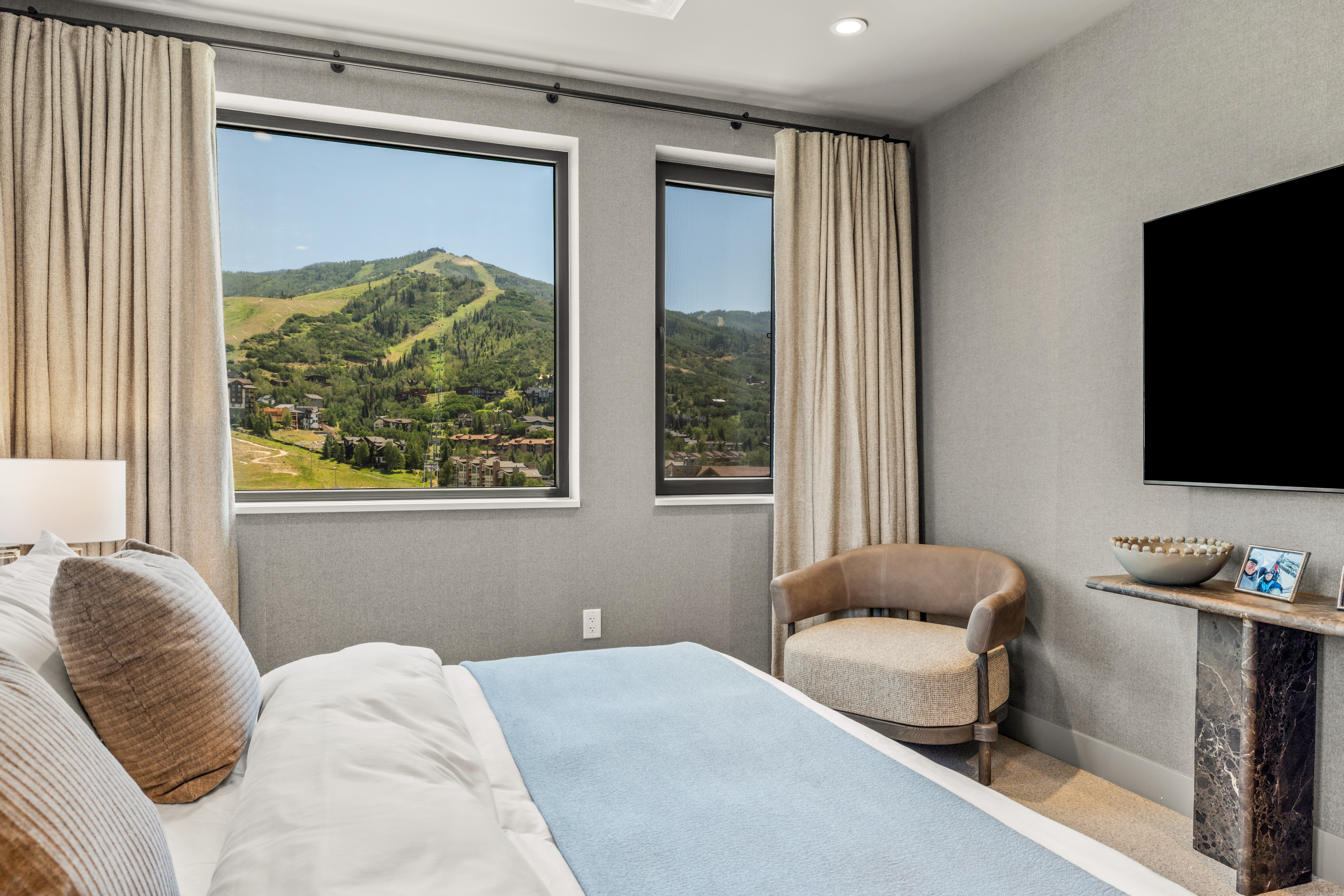 This bedroom features a neutral color palette with textured gray walls and a comfortable bed dressed in white linens with a light blue throw. Large windows provide a stunning, unobstructed view of a mountain ski resort, filling the room with natural light. A stylish, curved accent chair sits near the window, while a wall-mounted television is positioned above a sleek, dark stone console table.
