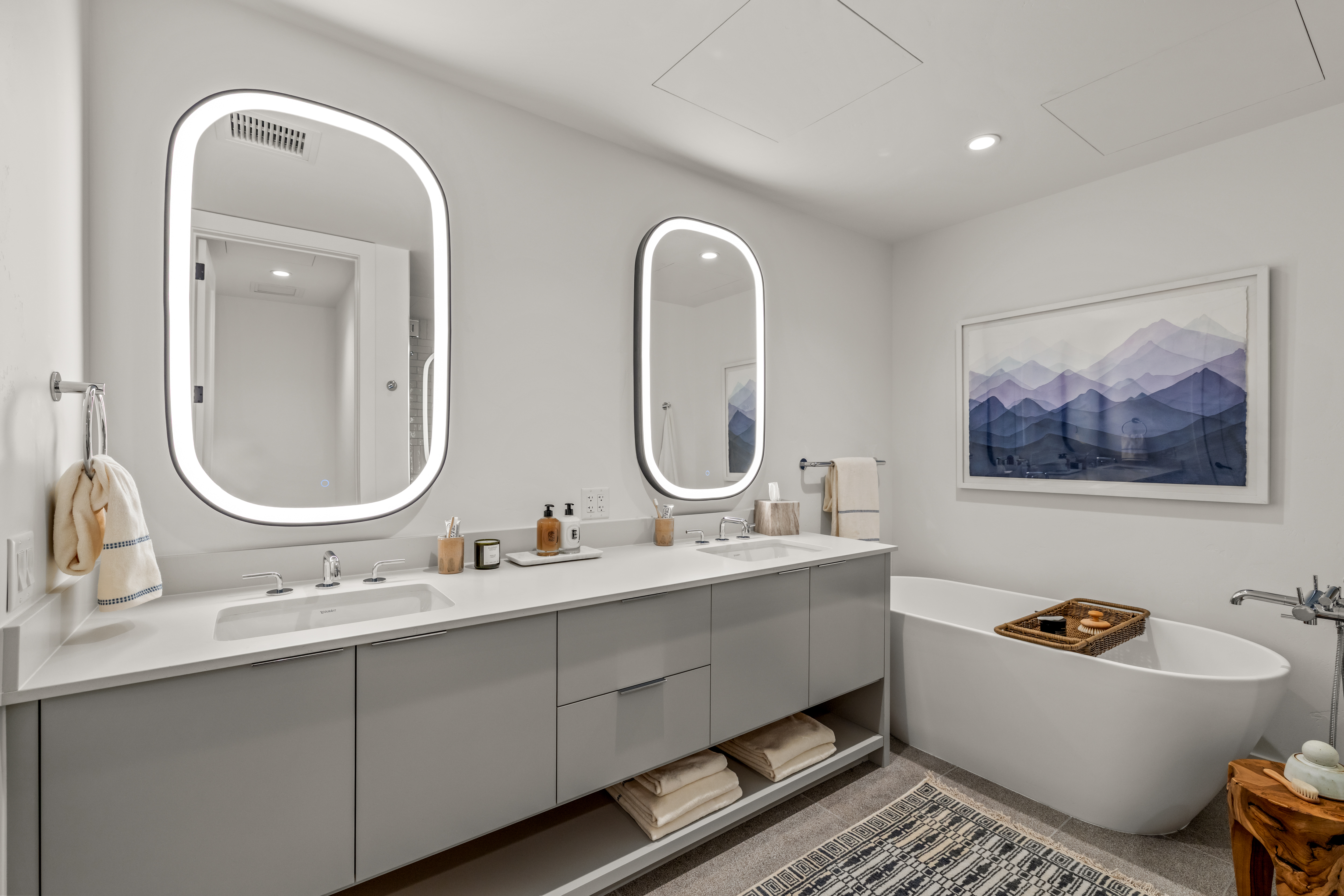This modern primary bathroom features a long, light gray dual-vanity with a white countertop and two illuminated, rounded-rectangular mirrors. To the right, a freestanding white soaking tub sits beneath a large, framed mountain-themed art piece, creating a serene spa-like atmosphere. The space is finished with neutral-toned floor tiles, a patterned area rug, and open shelving beneath the vanity for organized storage.