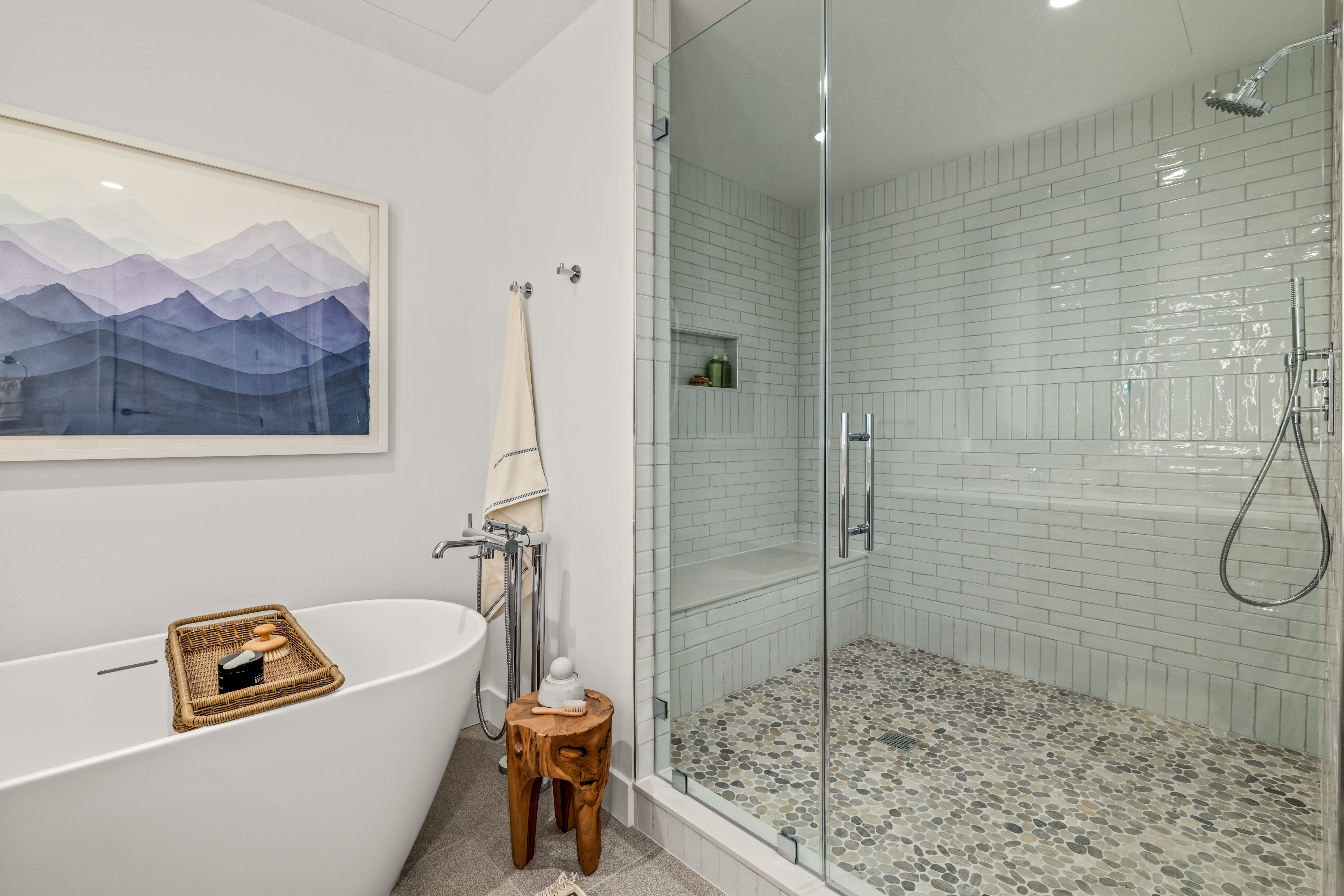 This modern primary bathroom features a sleek freestanding soaking tub paired with a spacious walk-in shower enclosed by glass panels. The shower is finished with white subway-style wall tiles and a textured pebble-stone floor, while a natural wood stool and a framed mountain landscape print add warmth to the space. The composition offers a clear, eye-level perspective that highlights the clean lines and spa-like atmosphere of the room.