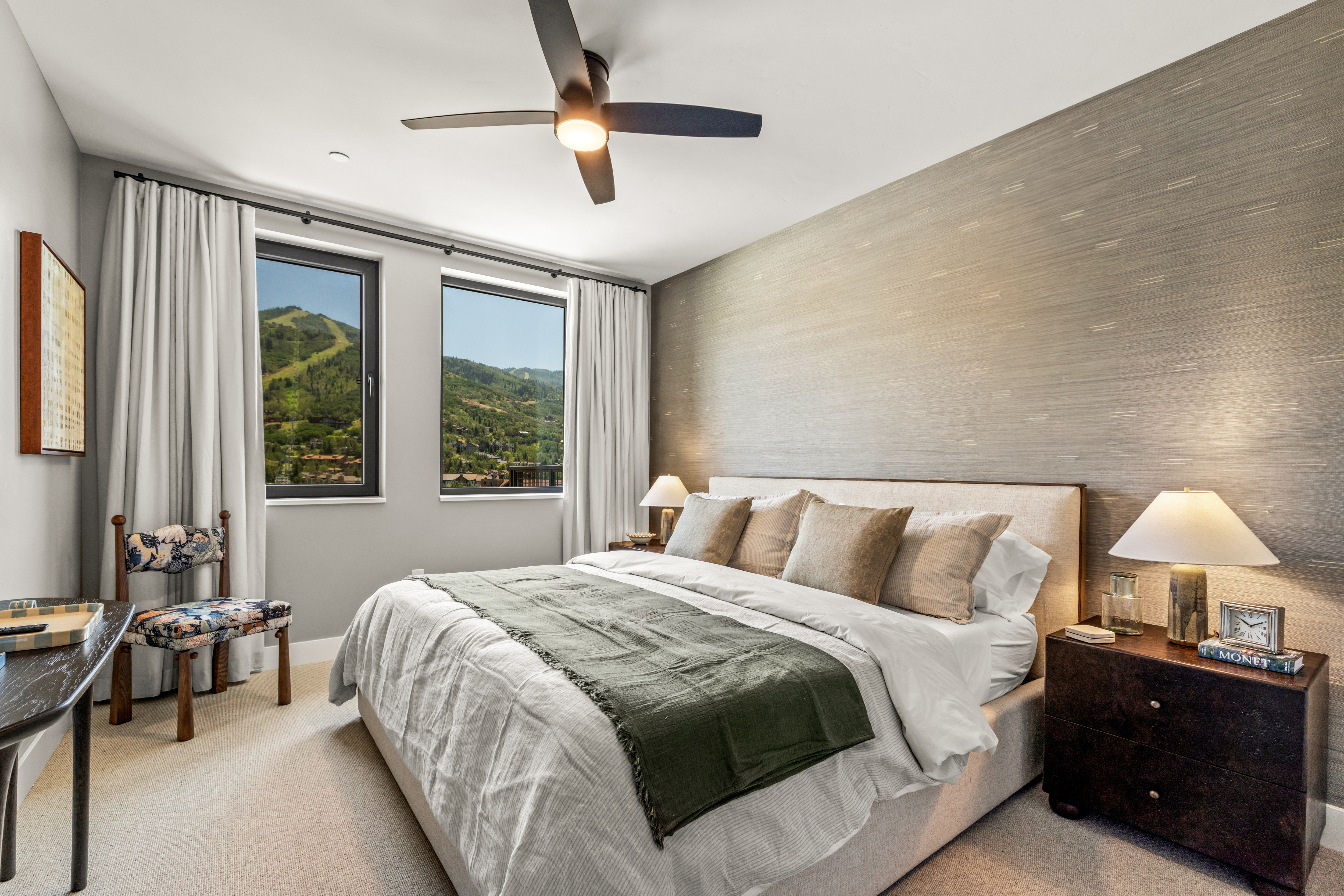 This serene bedroom features a neutral color palette with a textured accent wall behind a plush, upholstered bed. Large windows frame a scenic mountain view, while a patterned chair and dark wood nightstands add character to the space. The room is illuminated by a modern ceiling fan and warm bedside lamps, creating a cozy and inviting atmosphere.