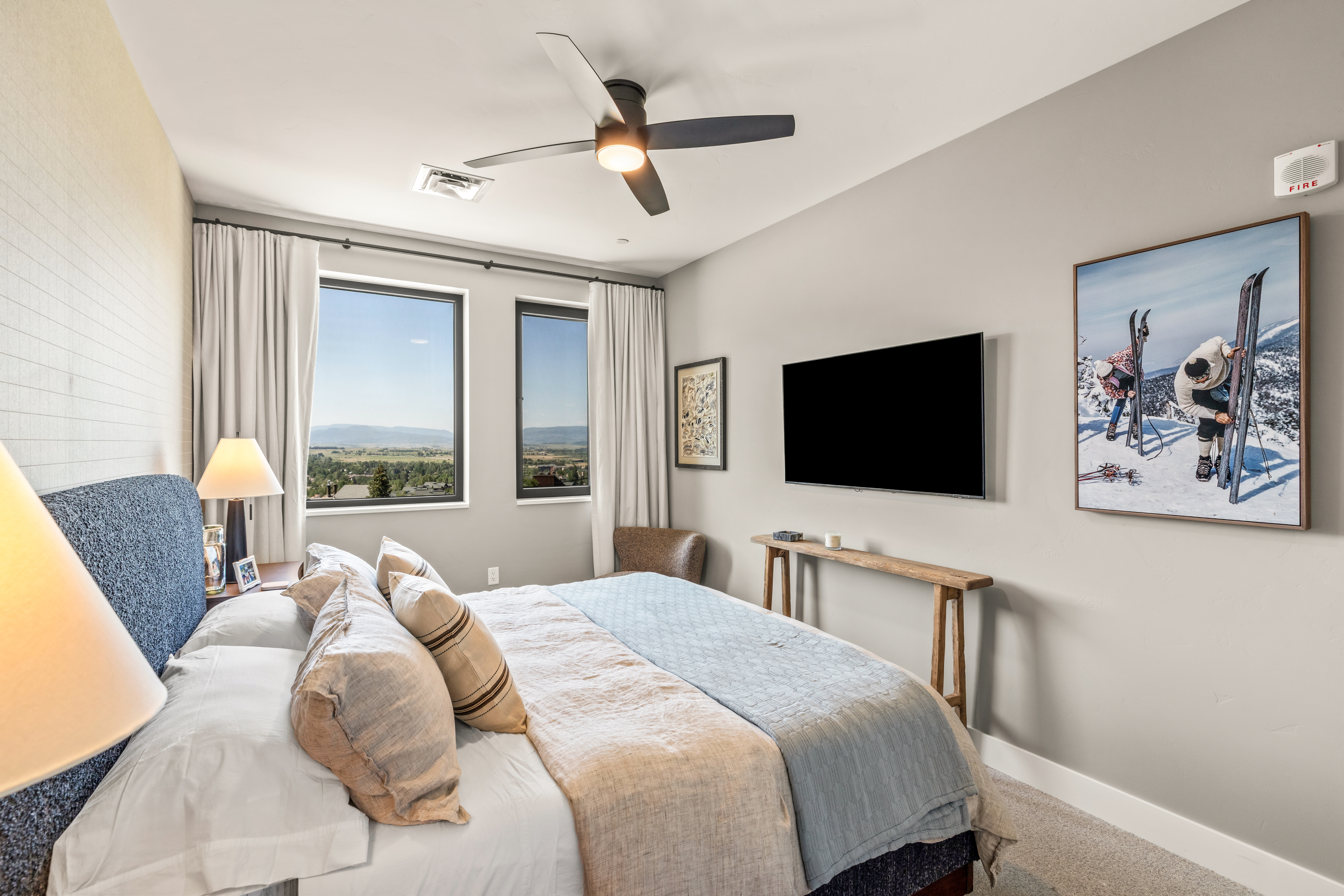 This well-appointed bedroom features a plush bed with a textured blue headboard and neutral bedding, positioned to take advantage of the scenic mountain views through two large windows. A wall-mounted television sits above a rustic wooden console table, while a large framed print of skiers adds a thematic touch to the space. The room is illuminated by a modern ceiling fan and a bedside lamp, creating a cozy and inviting atmosphere.