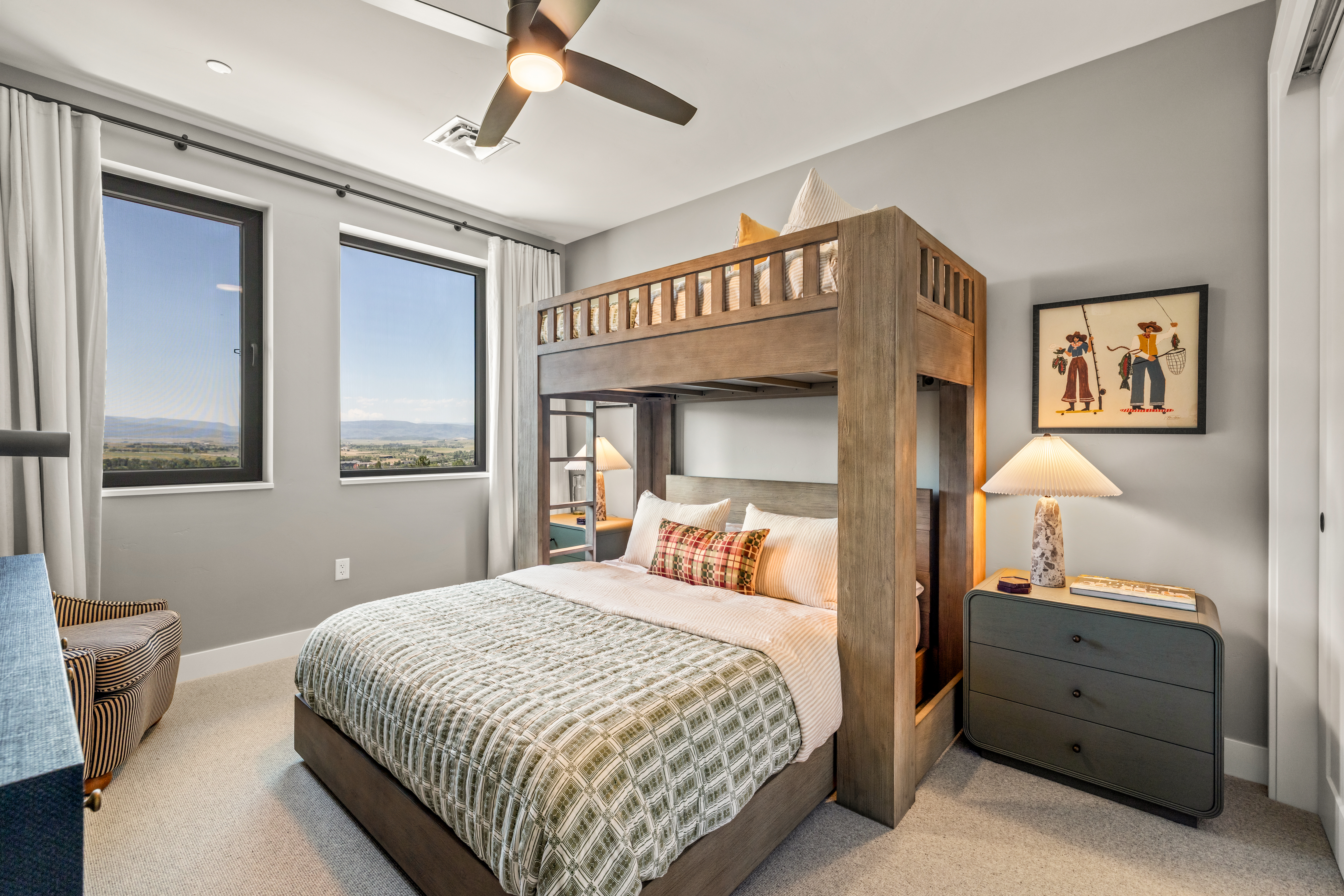 This bedroom features a unique wooden bunk bed structure that frames a lower bed, creating a cozy and functional sleeping area. The room is accented by a patterned quilt, a striped armchair, and a bedside table with a textured lamp, all set against neutral gray walls. Large windows provide ample natural light and a scenic view of the landscape beyond.