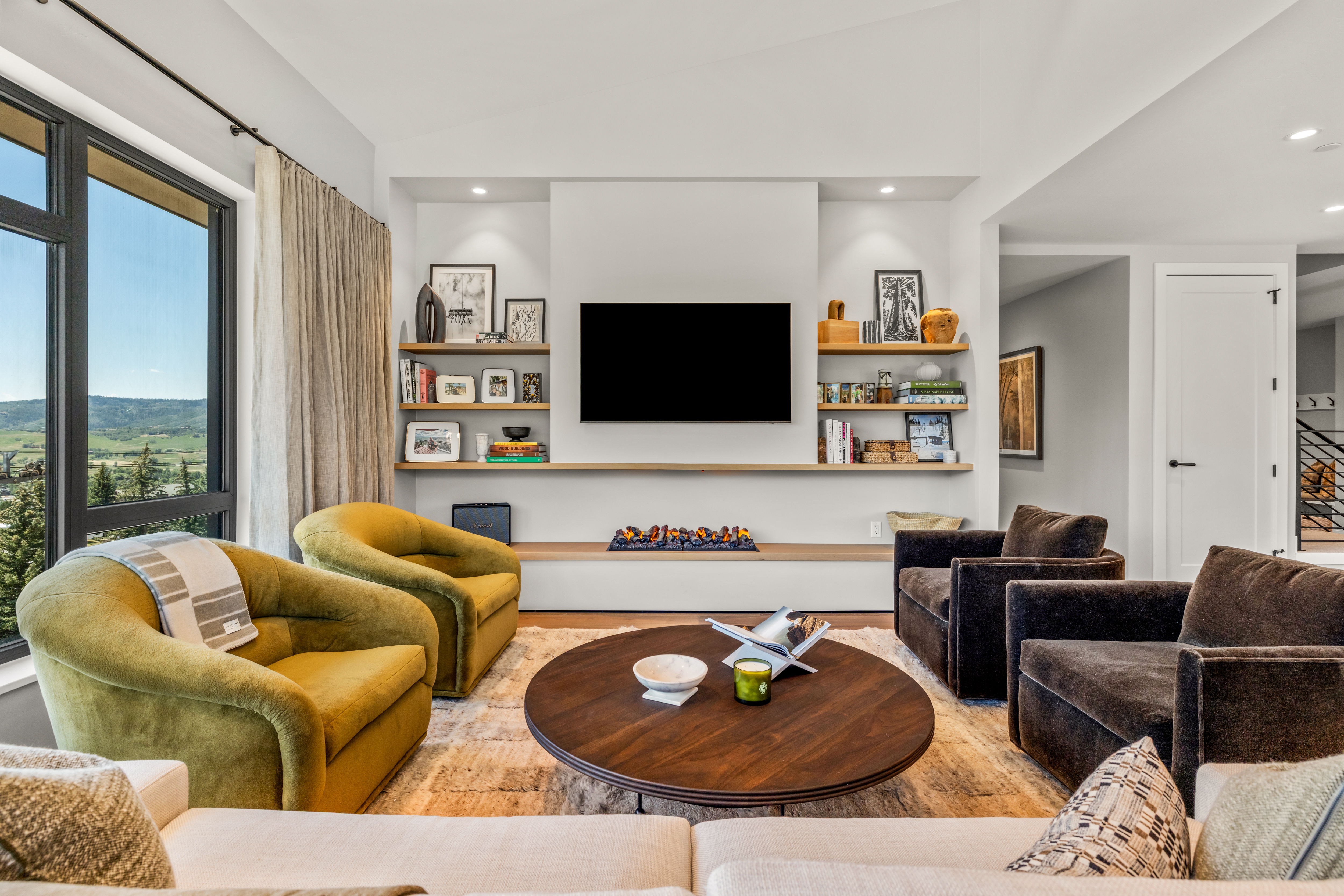 This sophisticated living room features a central media wall with a built-in television, open shelving, and a linear fireplace set into a long, low hearth. The space is furnished with a pair of mustard-colored armchairs, two dark velvet chairs, and a large circular coffee table resting on a textured area rug. Large windows to the left provide expansive views of the surrounding landscape, creating a bright and inviting atmosphere.