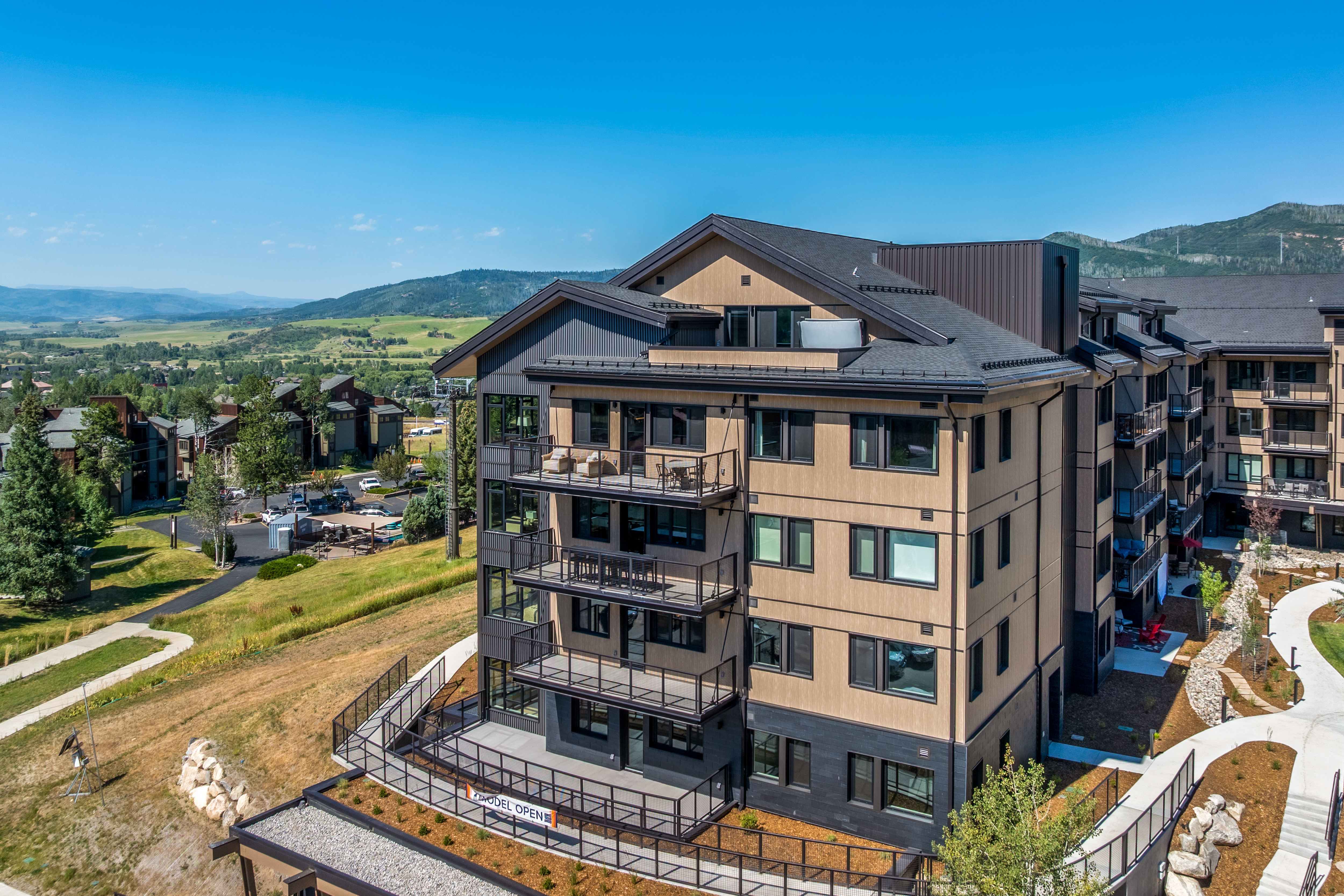 This high-angle aerial shot captures a modern, multi-story residential building set against a scenic mountainous landscape. The structure features a contemporary design with a mix of light wood-toned siding, dark metal accents, and multiple stacked balconies that offer expansive views. A paved walkway leads to the building's base, which is surrounded by landscaped grounds and a parking area, emphasizing a resort-like or mountain-community atmosphere.