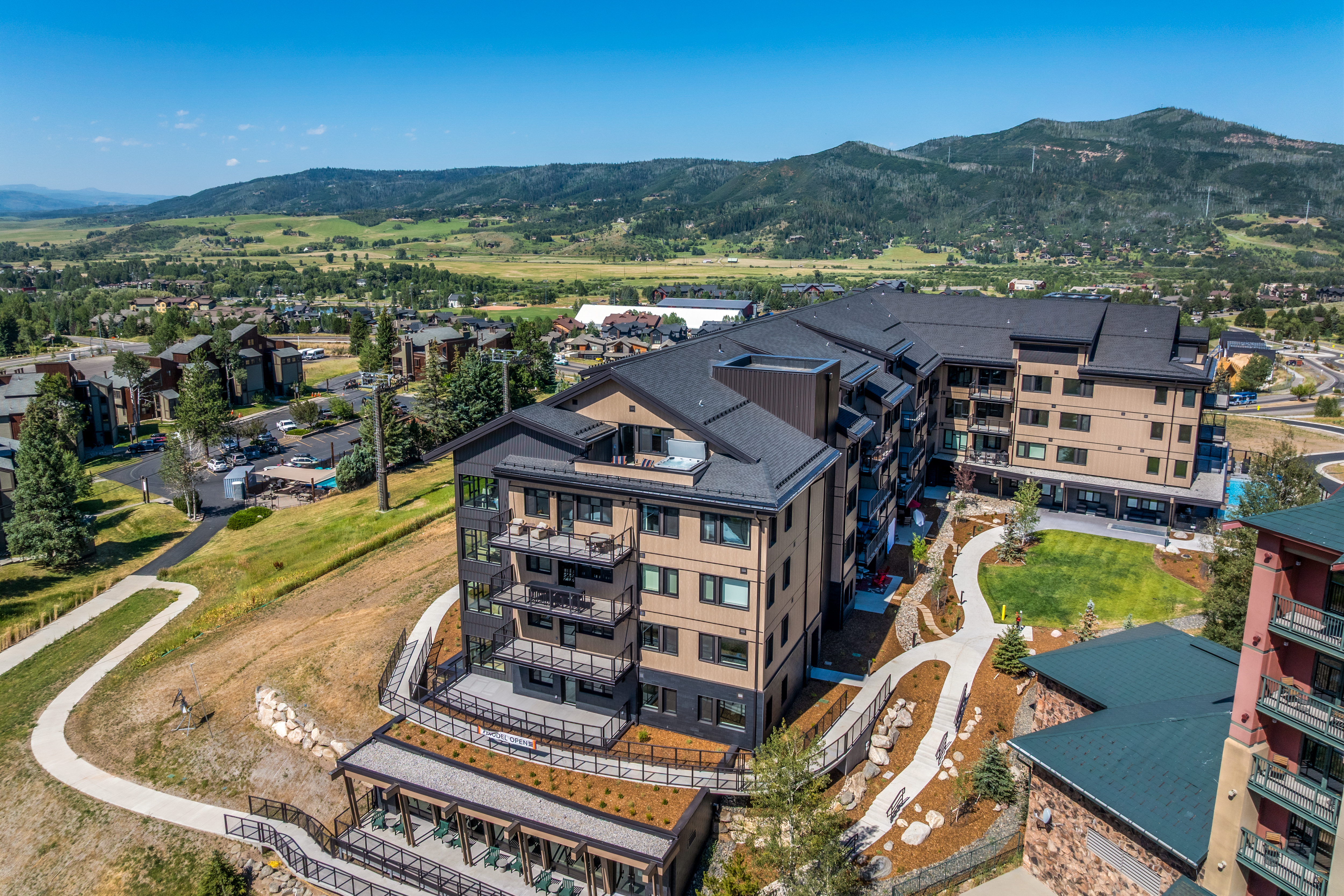 This high-angle aerial view captures a modern multi-story residential complex nestled in a scenic mountain landscape. The building features a contemporary design with a mix of wood-toned siding, dark metal accents, and multiple balconies, set against a backdrop of rolling green hills and distant peaks. A paved walkway winds through the landscaped grounds, connecting the structure to the surrounding community and natural terrain.