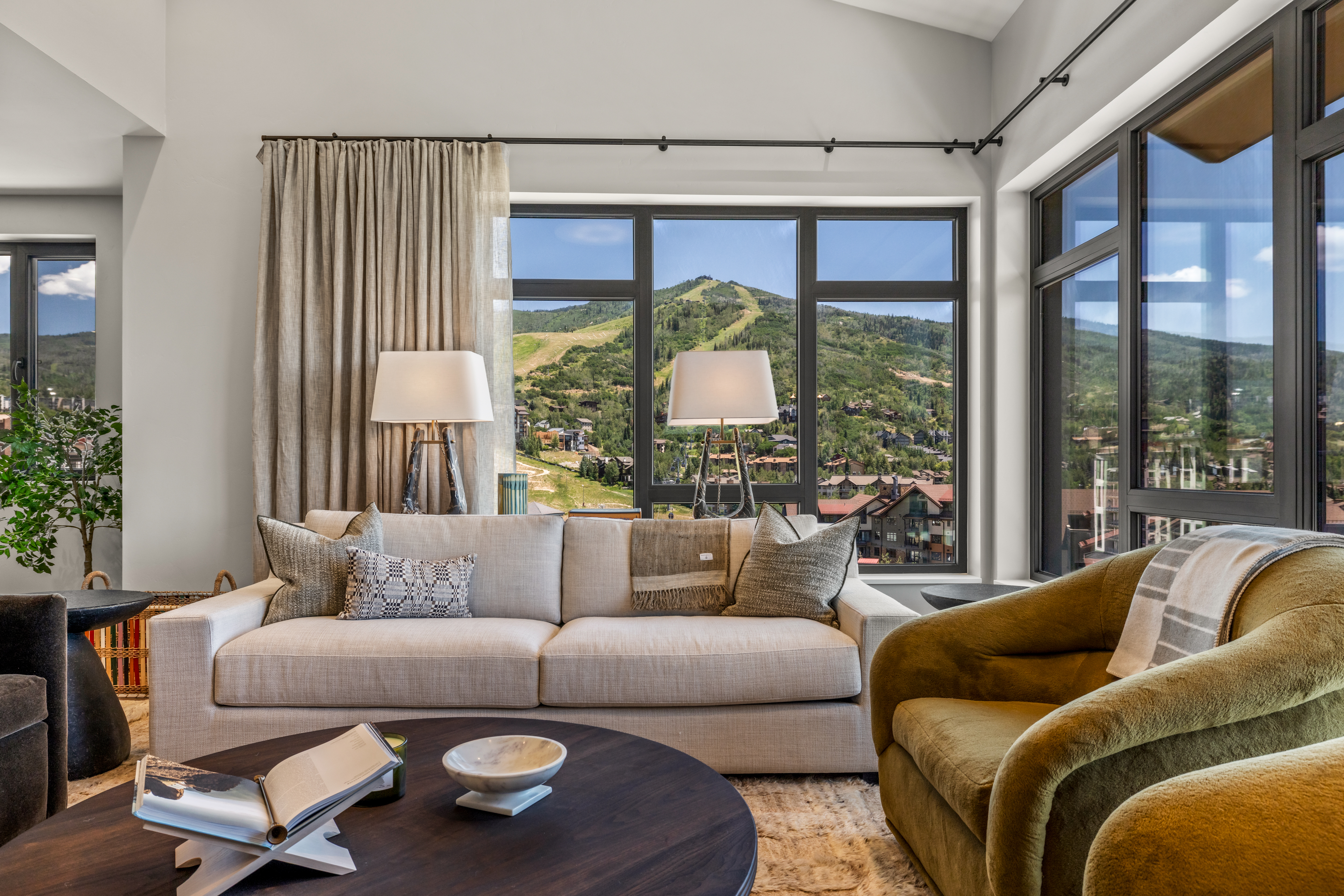 This inviting living room features a neutral-toned sofa accented with textured throw pillows and a draped blanket, positioned before large windows that frame a scenic mountain view. A dark, circular coffee table sits in the foreground, holding an open book and a decorative bowl, while a plush, olive-toned armchair adds a pop of color to the space. The room is illuminated by two matching table lamps, creating a warm and sophisticated atmosphere perfect for relaxing while enjoying the natural landscape.