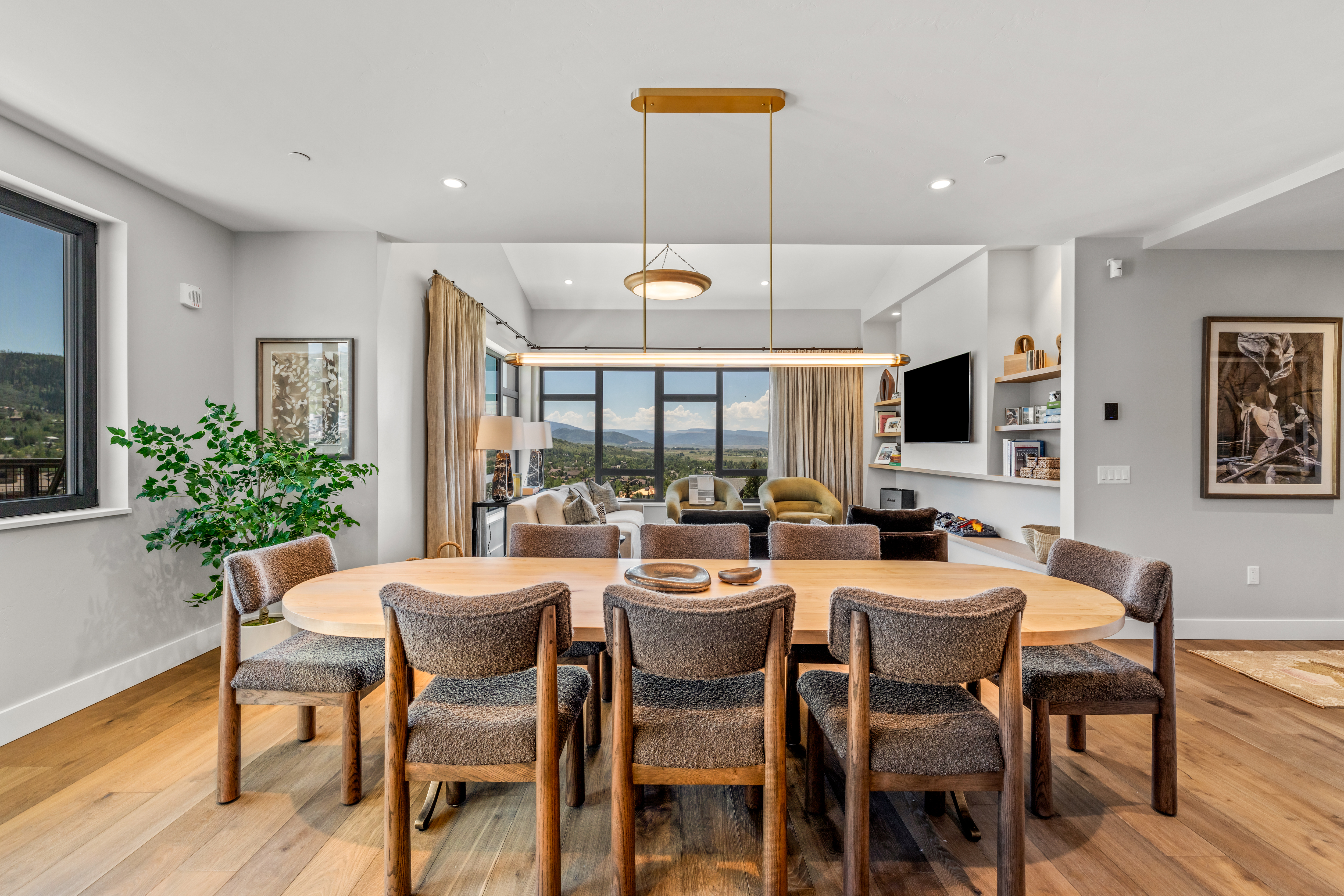 This elegant dining area features a large, light wood oval table surrounded by eight textured, dark-upholstered chairs, positioned beneath a modern linear pendant light. The room flows seamlessly into a living space with large windows that frame a scenic mountain view, while built-in shelving and a television are integrated into the adjacent wall. The overall aesthetic is sophisticated and contemporary, emphasizing an open-concept layout with warm, natural tones.