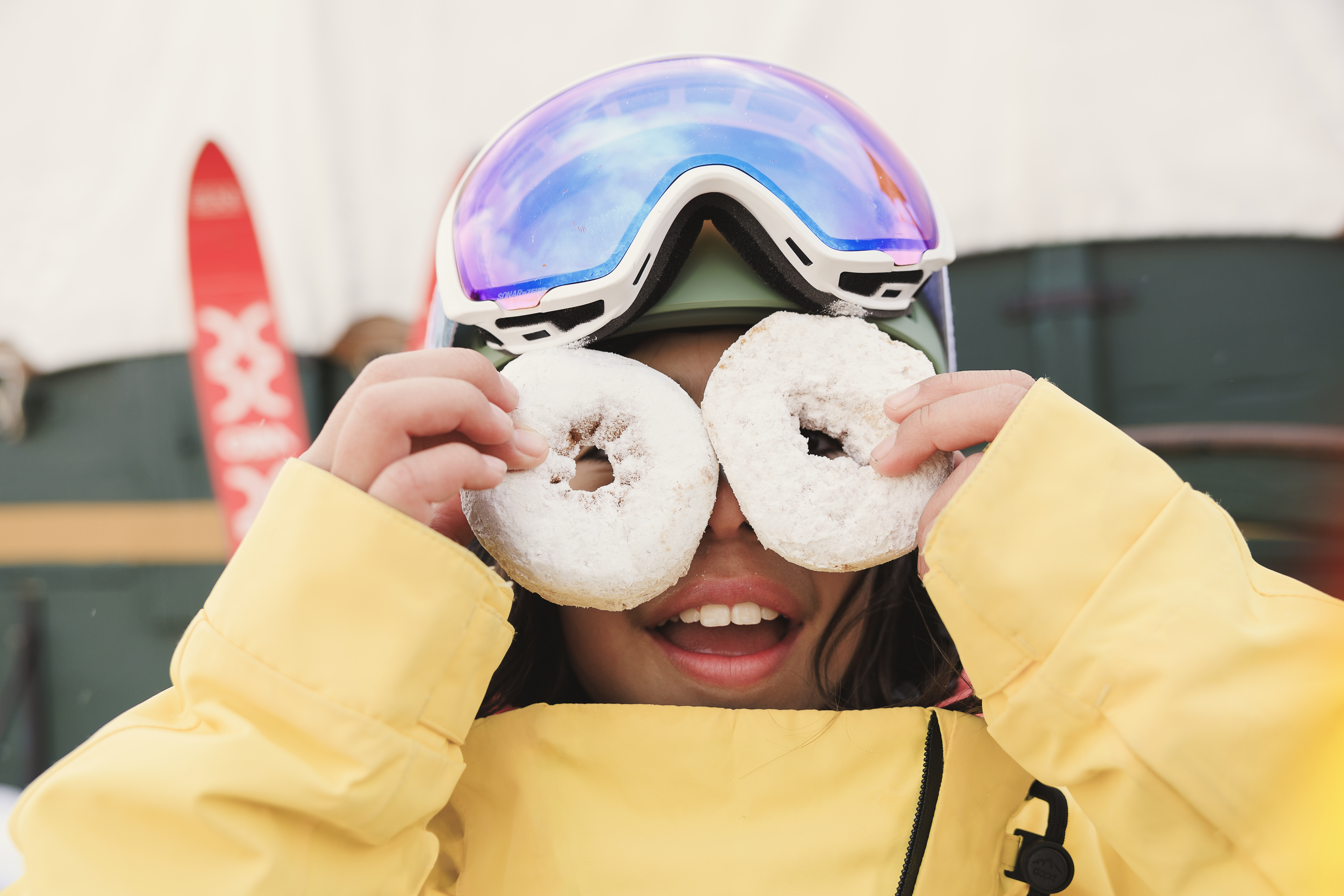 This close-up portrait features a person wearing a bright yellow winter jacket and a ski helmet with colorful goggles pushed up onto the forehead. The individual is playfully holding two powdered donuts in front of their eyes, creating a whimsical and lighthearted scene. The background shows a blurred outdoor setting with a red ski visible, suggesting a recreational or mountain resort environment.