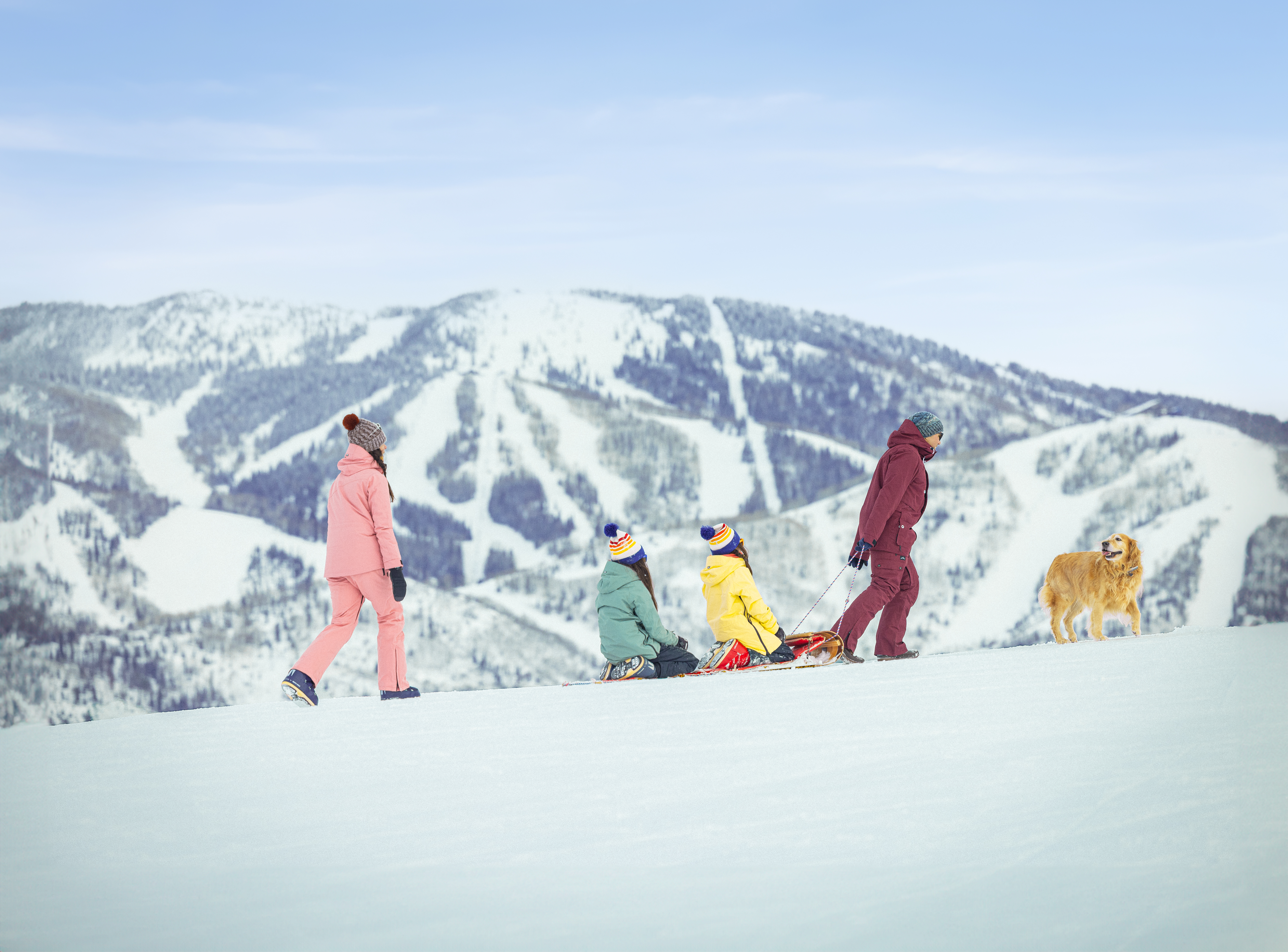 This scenic outdoor shot captures a family enjoying a winter day on a snowy slope with a large mountain resort in the background. The perspective is a wide, eye-level cinematic shot that emphasizes the vast, open landscape and the recreational lifestyle associated with the property's location. The clear blue sky and expansive snow-covered terrain highlight the prime positioning of the community near world-class skiing facilities.