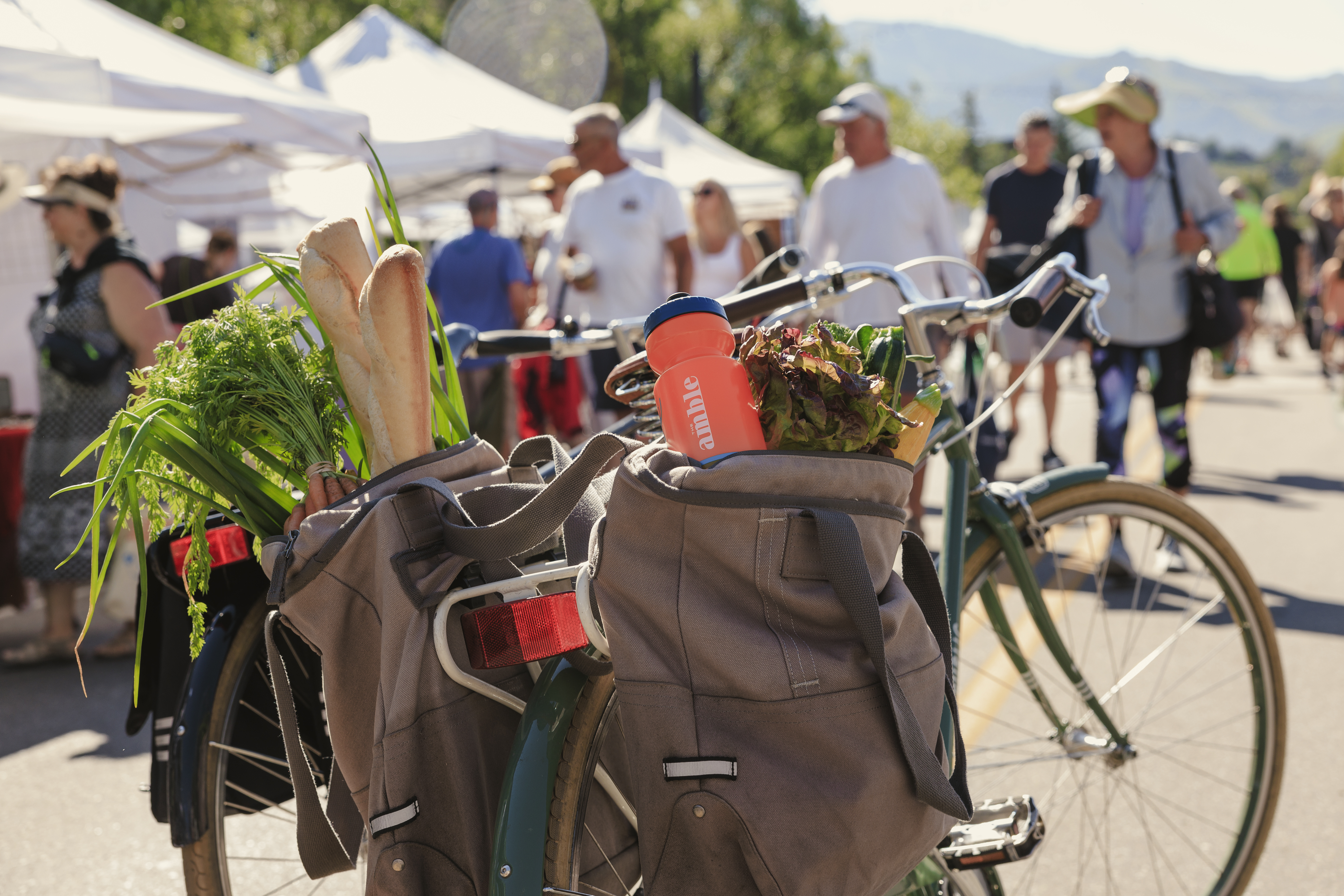 This close-up, eye-level shot captures a bicycle parked at a bustling outdoor farmers market, with its pannier bags overflowing with fresh produce like carrots, baguettes, and leafy greens. An orange water bottle branded with 'amble' sits prominently in one of the bags, while a blurred crowd of shoppers and white vendor tents fill the background under a bright, sunny sky. The scene evokes a vibrant, active community lifestyle, highlighting the convenience of local amenities and outdoor recreation.