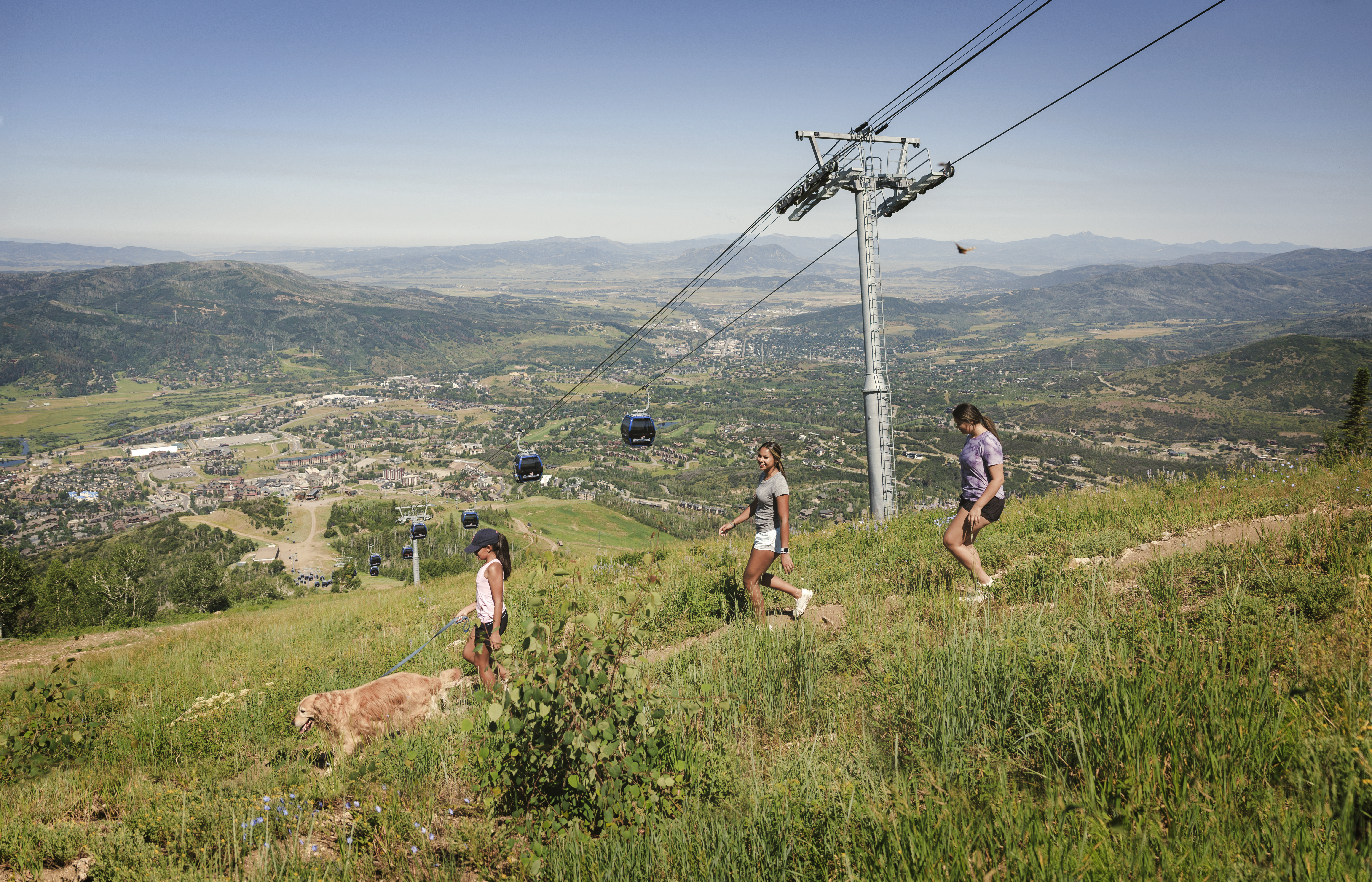 This scenic outdoor shot captures a hillside trail overlooking a sprawling mountain town and valley. Three individuals and a dog are walking along the grassy path, positioned near a large ski lift tower and gondola cables that stretch across the landscape. The perspective is wide and elevated, emphasizing the expansive recreational access and natural beauty of the surrounding community.