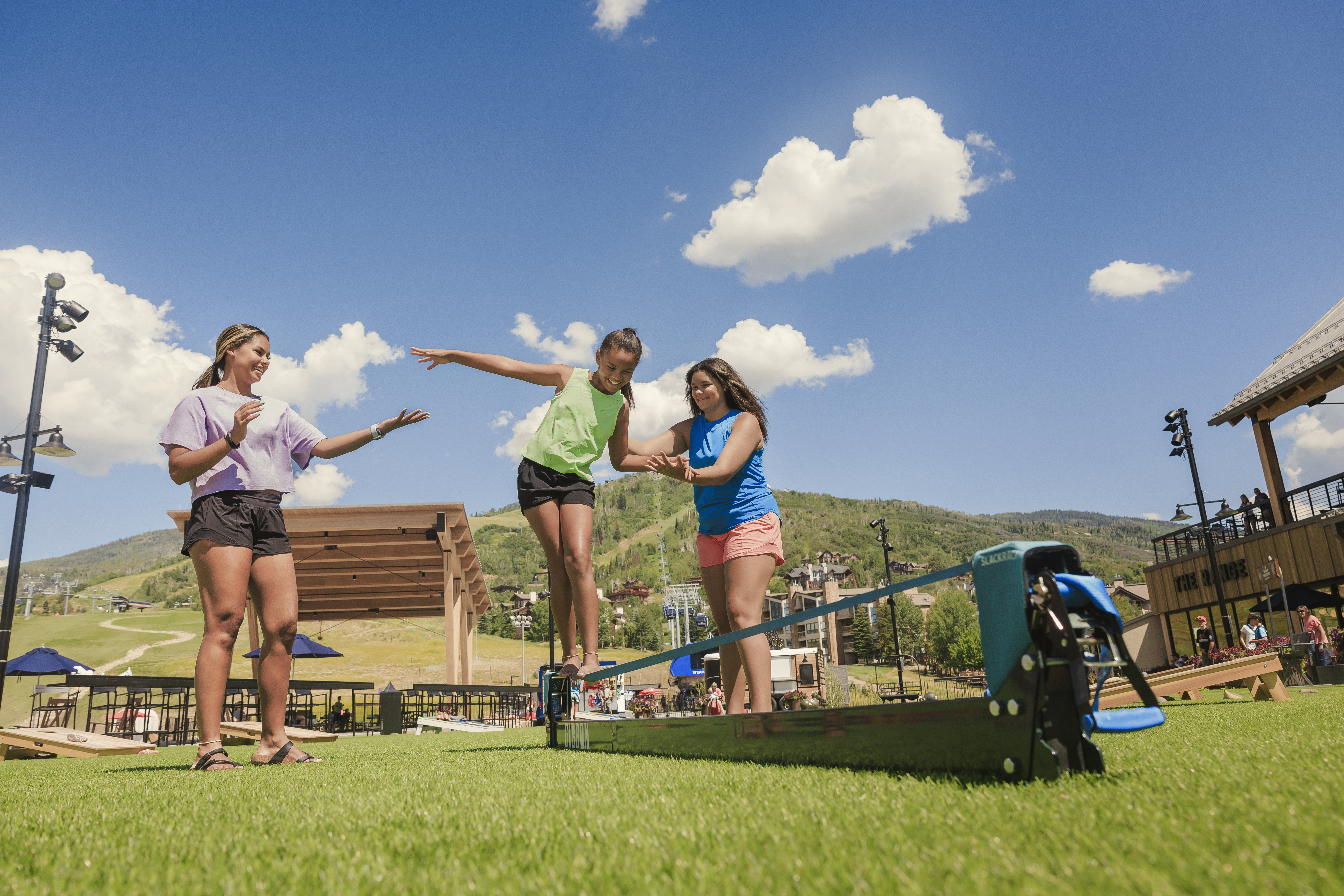 This outdoor recreational area features a slackline setup on a manicured lawn, with three individuals engaging in a balancing activity. The background showcases a mountain resort setting with a wooden pavilion, a multi-story building, and rolling hills under a bright, clear blue sky. The scene captures a vibrant, active lifestyle common in mountain community amenities.