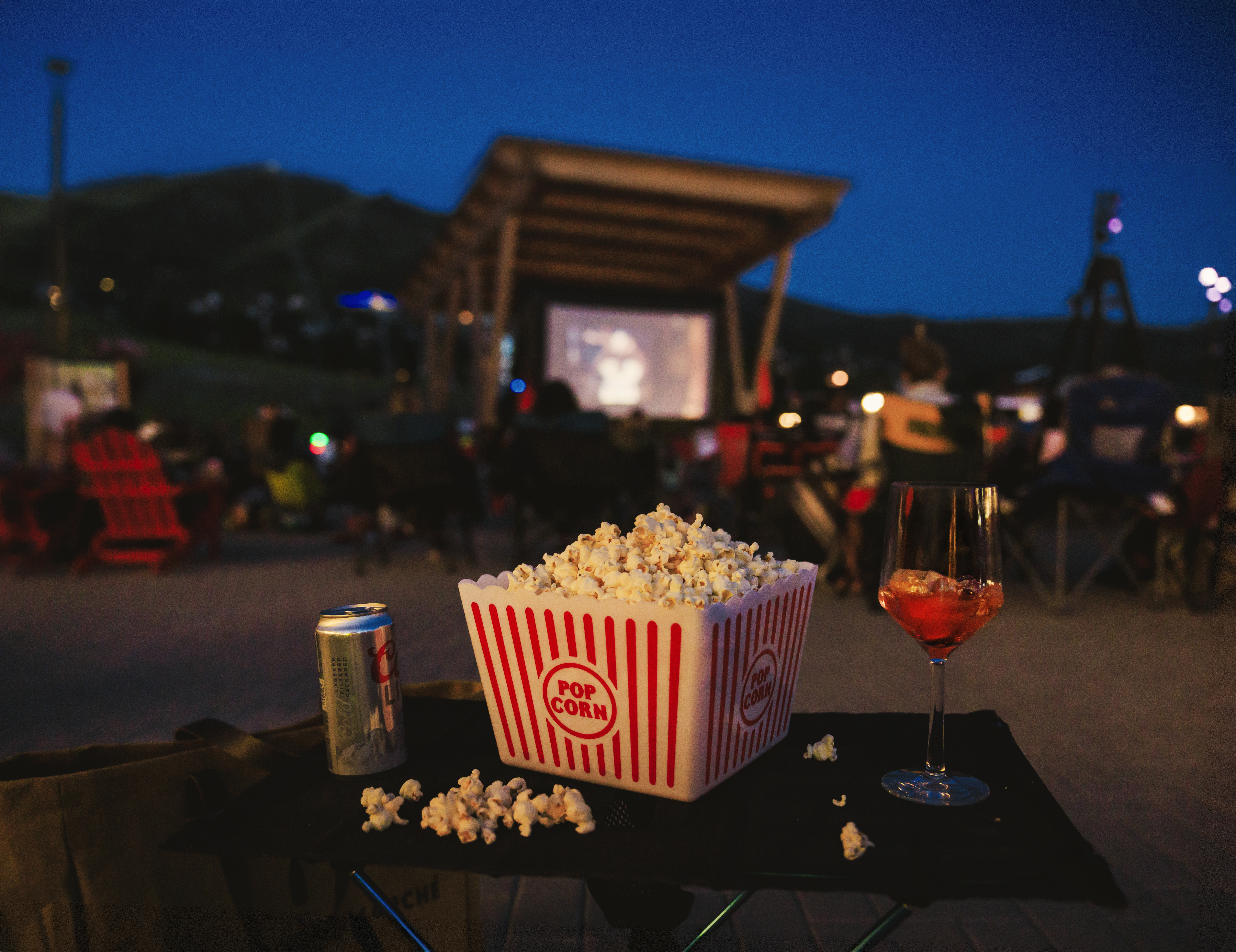 This image captures an outdoor movie night setting, featuring a foreground table with a bucket of popcorn, a drink, and a glass of wine. In the background, a large projection screen is set up under a covered structure, with audience members seated in chairs on a paved area. The scene is set during twilight, creating a relaxed and inviting community atmosphere.