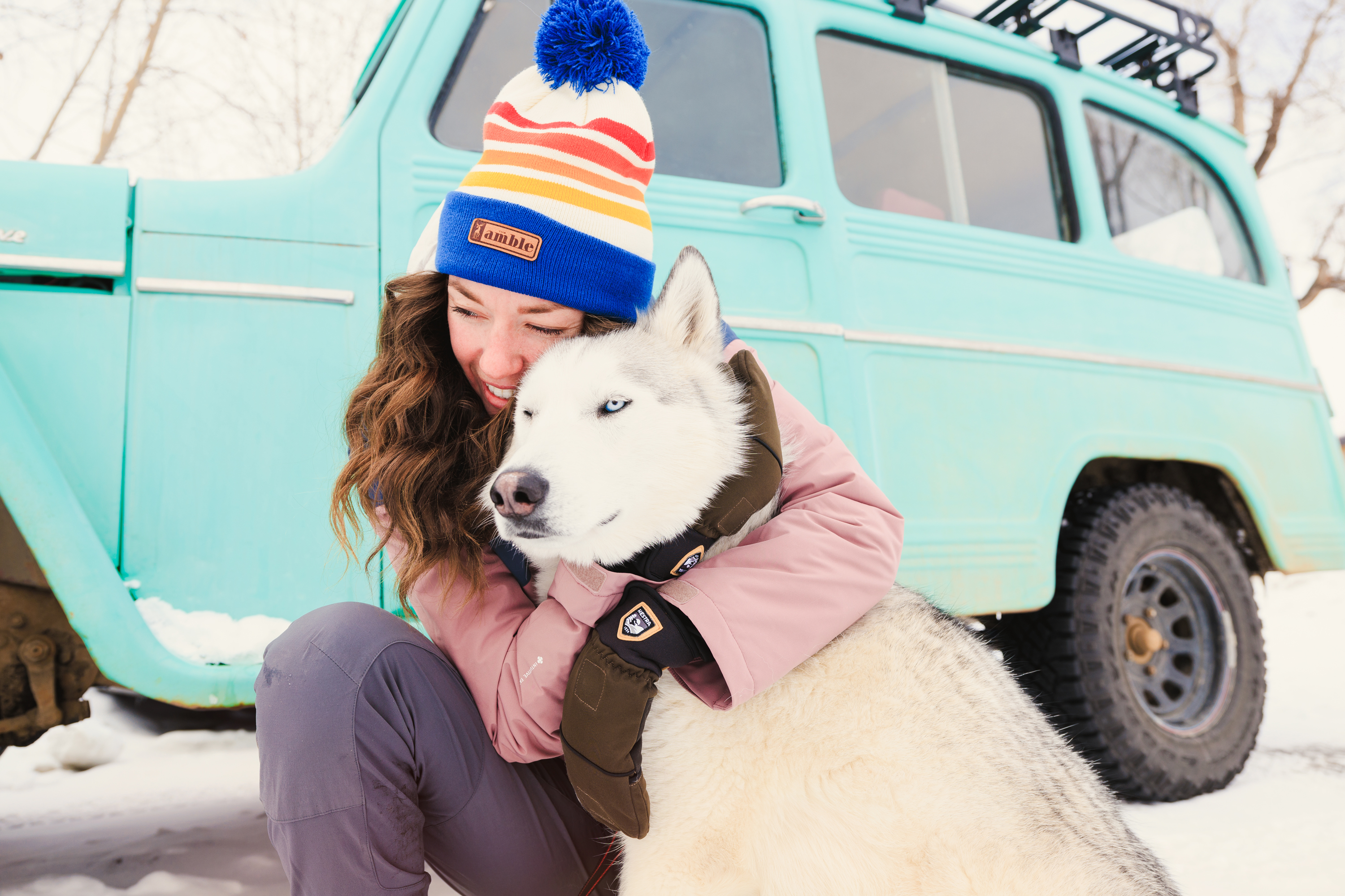 A woman wearing a colorful knit beanie and a pink jacket is kneeling in the snow, embracing a white husky. In the background, a vintage, light blue station wagon with a roof rack is parked on a snowy surface. The scene captures a cozy, outdoor winter lifestyle aesthetic with a focus on the interaction between the person and the dog.