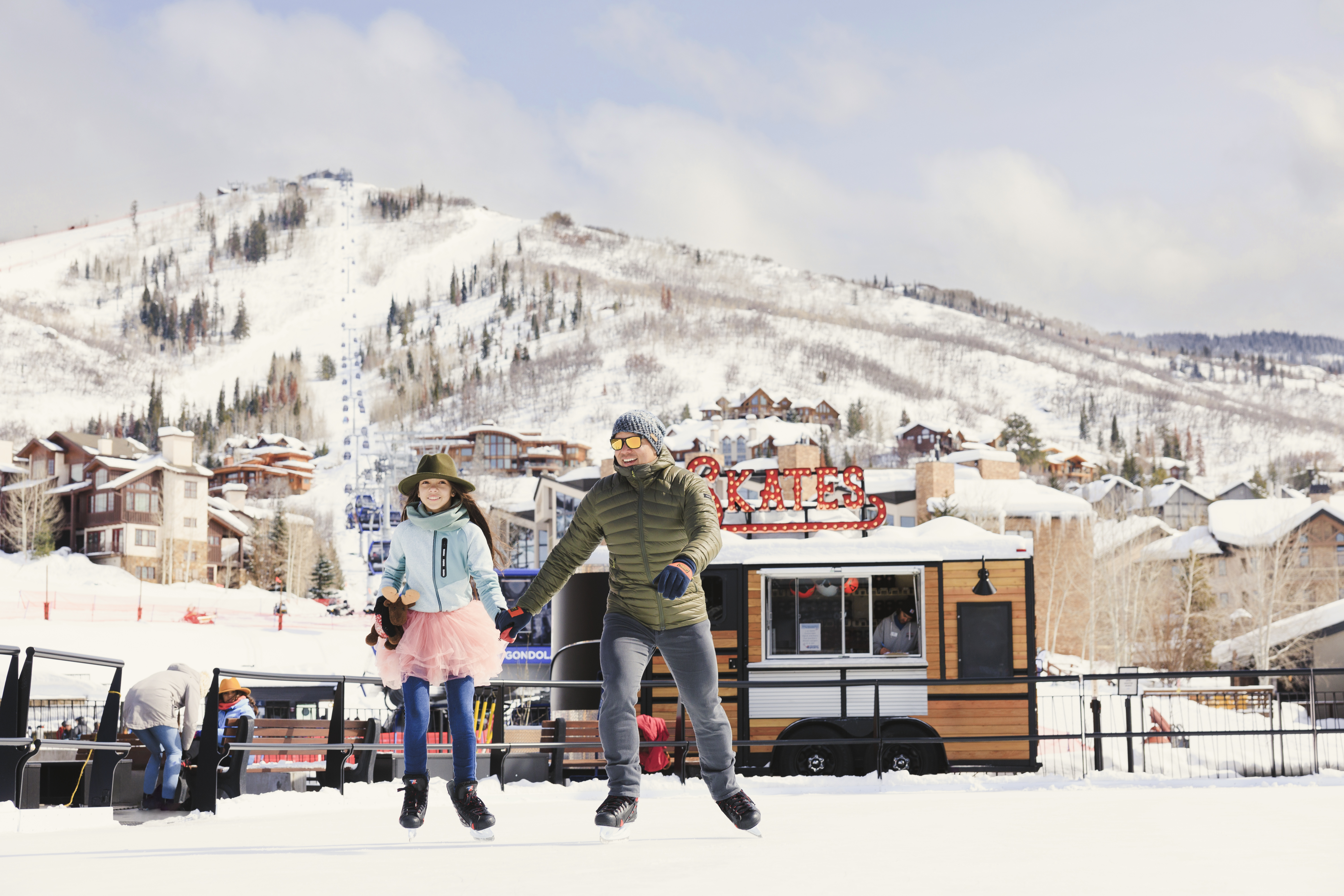 This image captures a vibrant outdoor ice skating rink set against a scenic mountain backdrop, highlighting a desirable community amenity. A man and a young girl are skating in the foreground, while a rustic-style mobile skate rental kiosk with a prominent 'SKATES' sign sits nearby. The scene conveys an active, family-friendly lifestyle with snow-covered slopes and residential buildings visible in the distance.