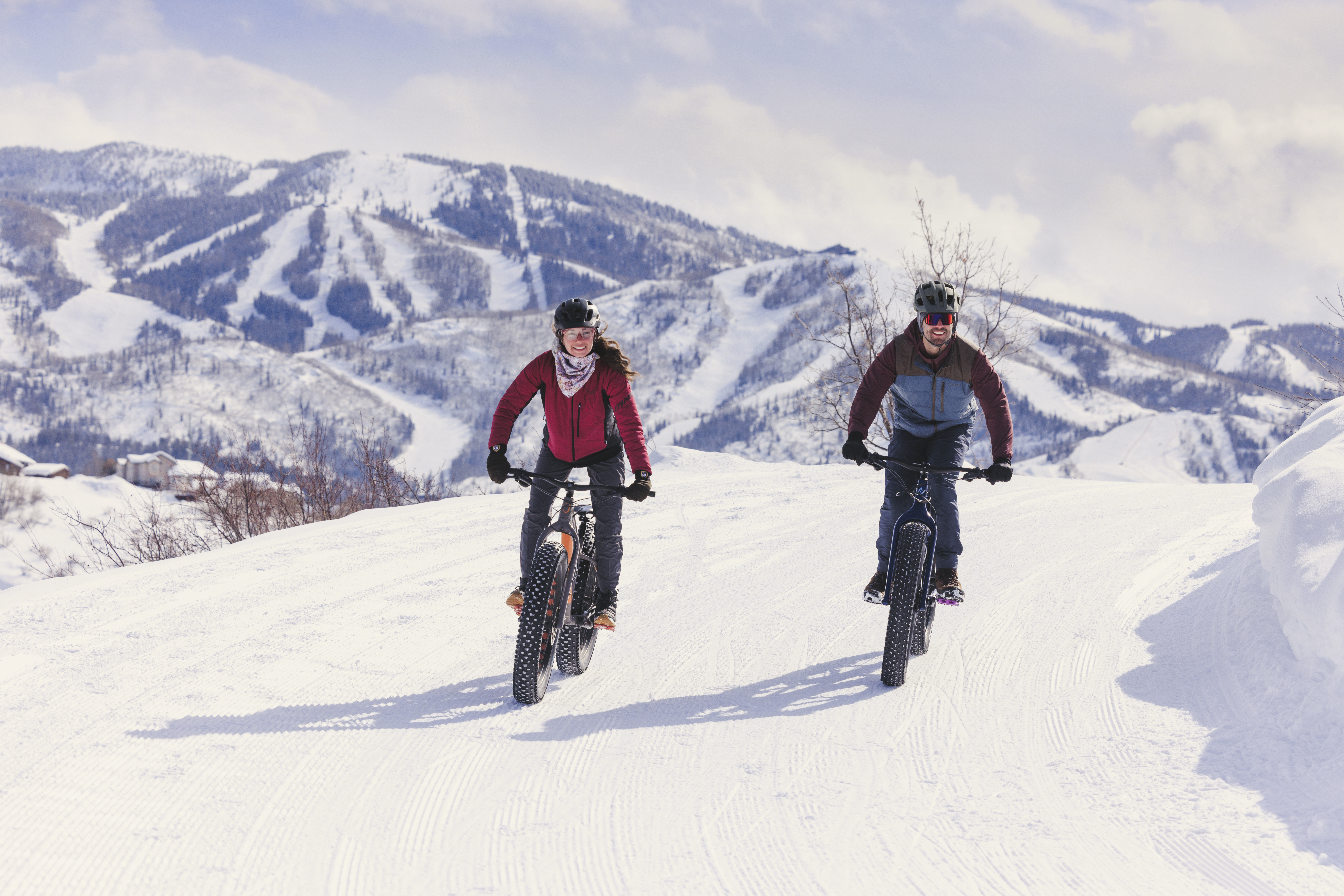 This image captures two people riding fat-tire bikes across a groomed snow-covered trail, highlighting the outdoor recreational lifestyle available in the community. In the background, a sprawling mountain resort with ski runs and residential structures is visible under a bright, partly cloudy sky. The perspective is a wide, eye-level shot that emphasizes the expansive winter landscape and the active, adventurous nature of the location.