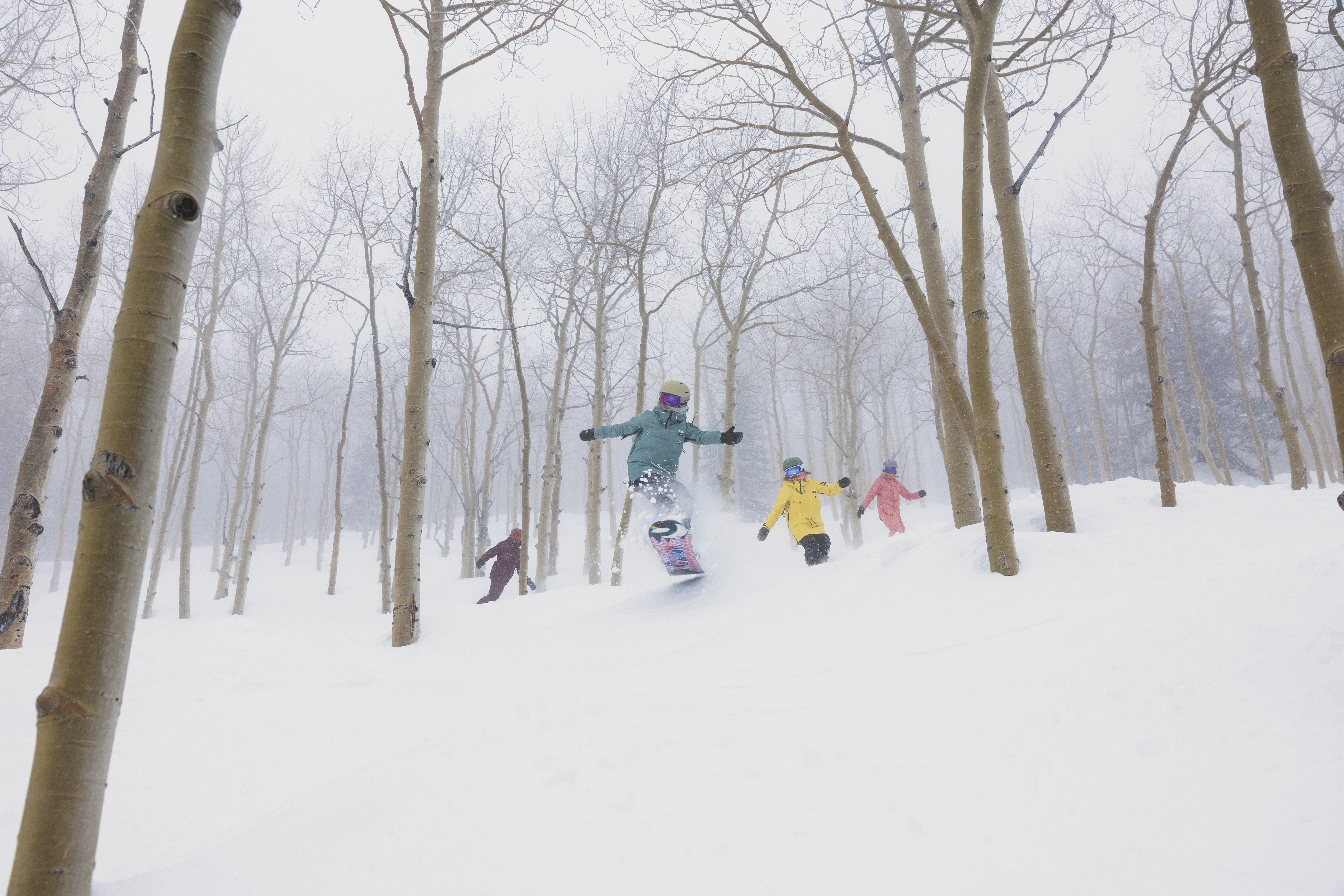 This action-oriented outdoor scene captures a group of people snowboarding through a dense, snow-covered forest of aspen trees. The perspective is at eye-level, emphasizing the dynamic movement of the central snowboarder mid-air against a backdrop of misty, winter woods. The atmosphere is cold and adventurous, highlighting the recreational potential of a mountainous, snowy landscape.