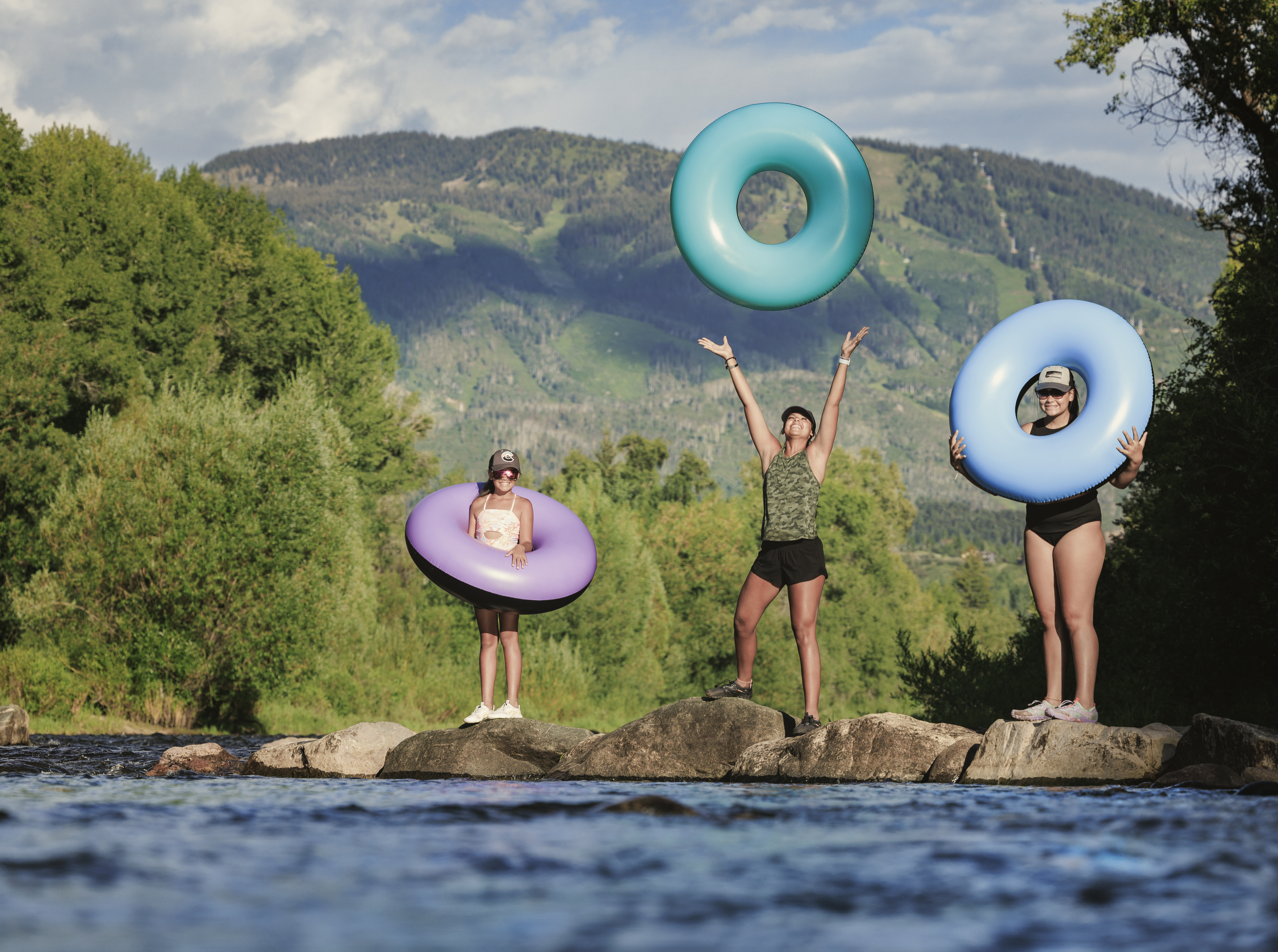 This image captures three people standing on rocks in a river, holding colorful inflatable tubes against a backdrop of lush green trees and a large mountain range. The scene is shot from a low, eye-level perspective, emphasizing the outdoor recreational lifestyle and natural beauty of the surrounding landscape. The bright, natural lighting highlights the vibrant colors of the tubes and the scenic mountain environment.