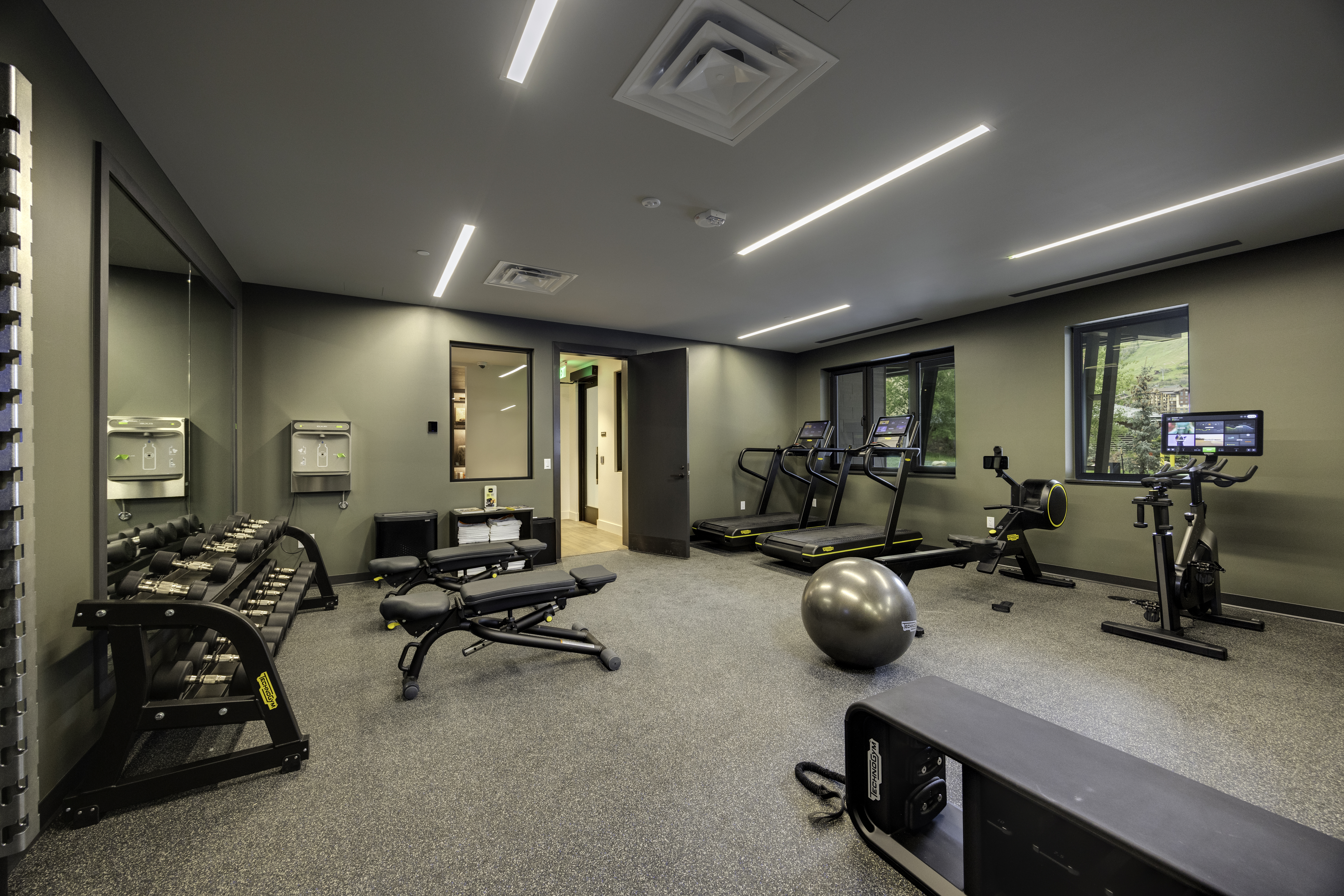 This well-equipped home gym features a professional layout with a variety of Technogym equipment, including treadmills, a rowing machine, and a stationary bike. The space is finished with durable, speckled grey flooring, olive-toned walls, and recessed linear lighting, creating a clean and functional workout environment. A large wall mirror enhances the sense of space, while windows provide natural light and views of the outdoors.