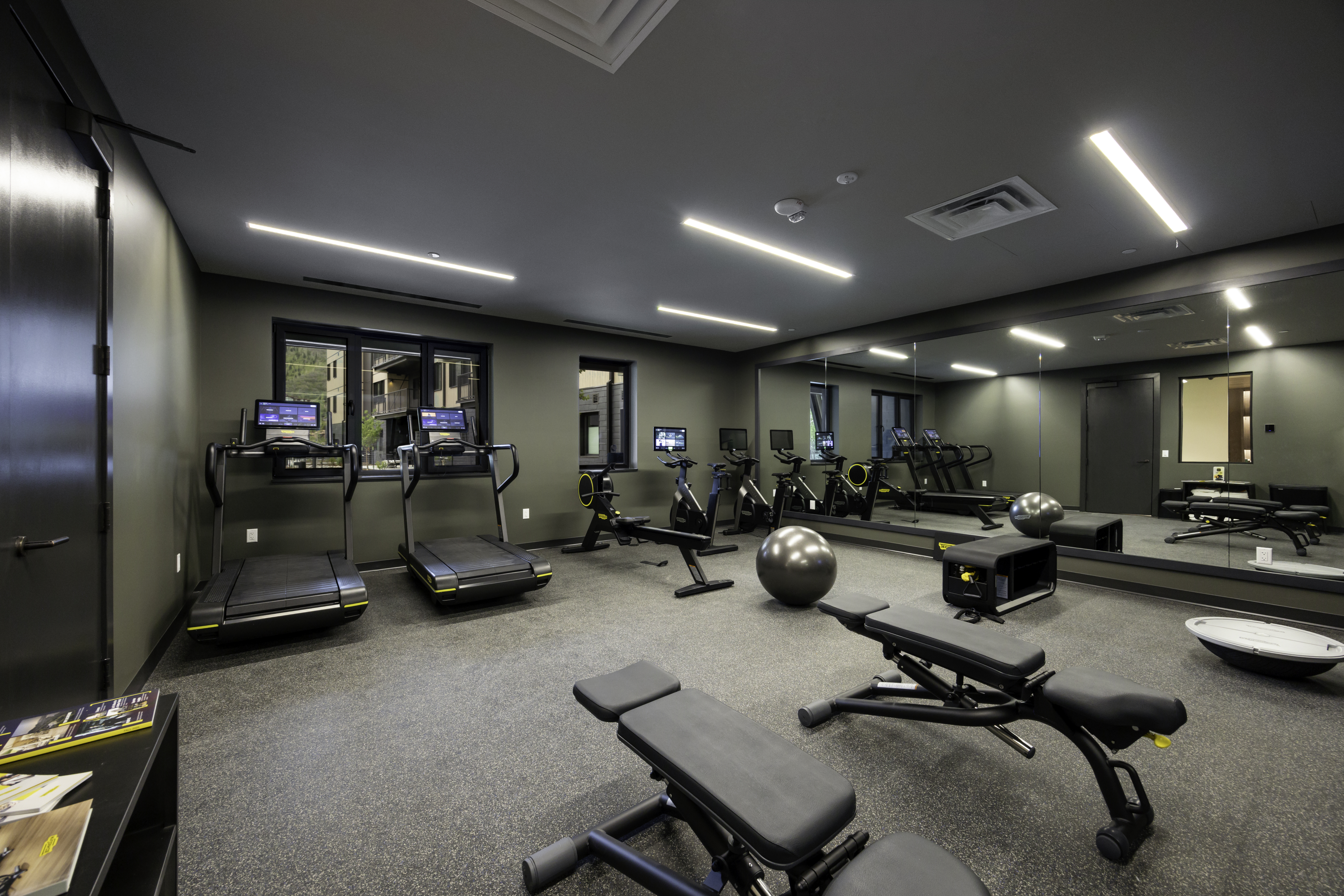 This modern, well-equipped home gym features a dark, minimalist aesthetic with olive-toned walls and durable grey flooring. The space is outfitted with two treadmills, a rowing machine, an exercise bike, and multiple adjustable weight benches, all arranged for optimal flow. A large wall-mounted mirror expands the room's visual depth, reflecting the equipment and the bright, linear LED lighting overhead.