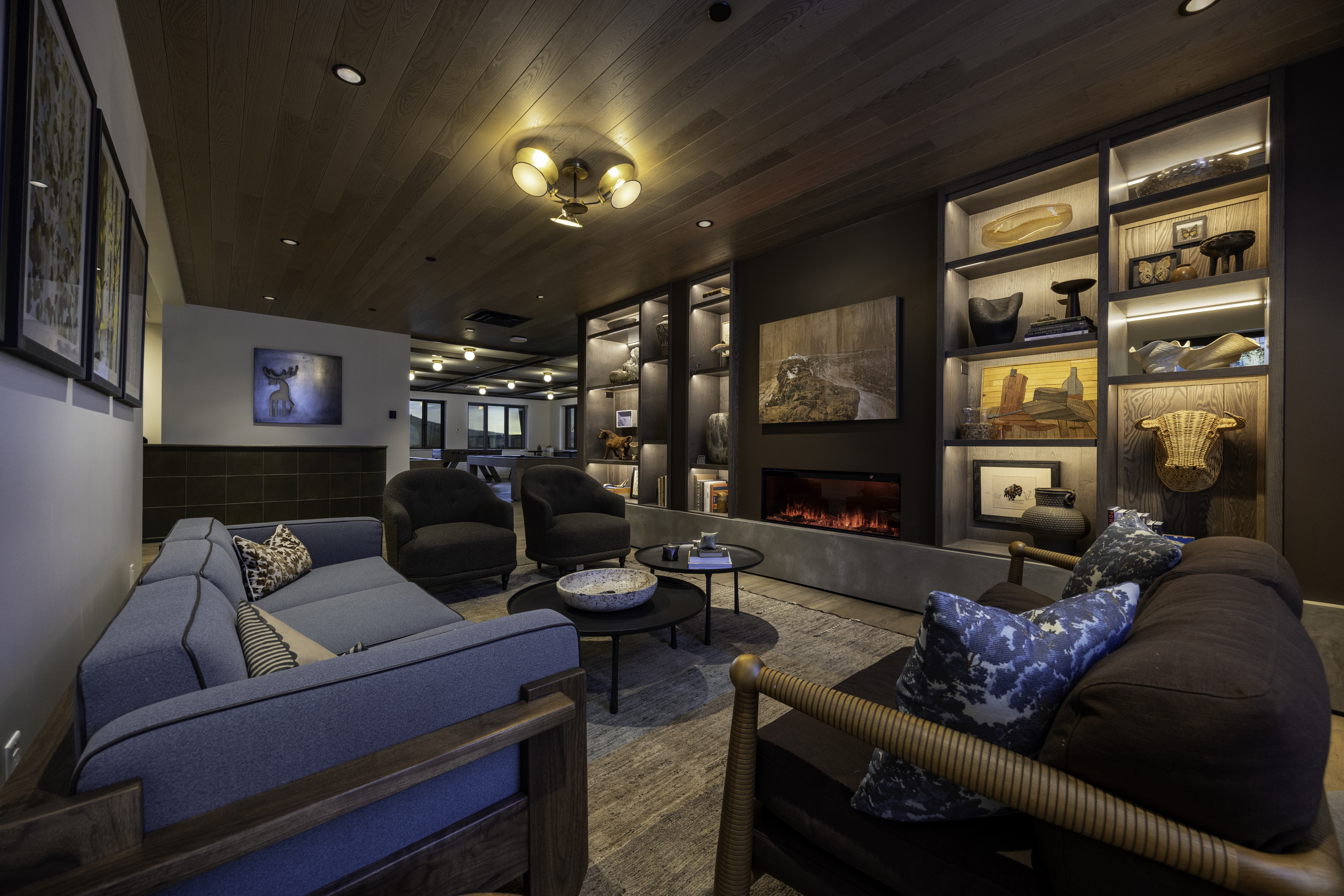 This sophisticated living room features a warm, inviting atmosphere with a wood-paneled ceiling and a central fireplace integrated into a dark, built-in shelving unit. The space is furnished with a blue sofa, two dark armchairs, and a secondary brown sofa, all arranged around two circular coffee tables on a textured area rug. The room exudes a cozy, lodge-inspired aesthetic, with recessed lighting and decorative art pieces displayed on the illuminated shelves.