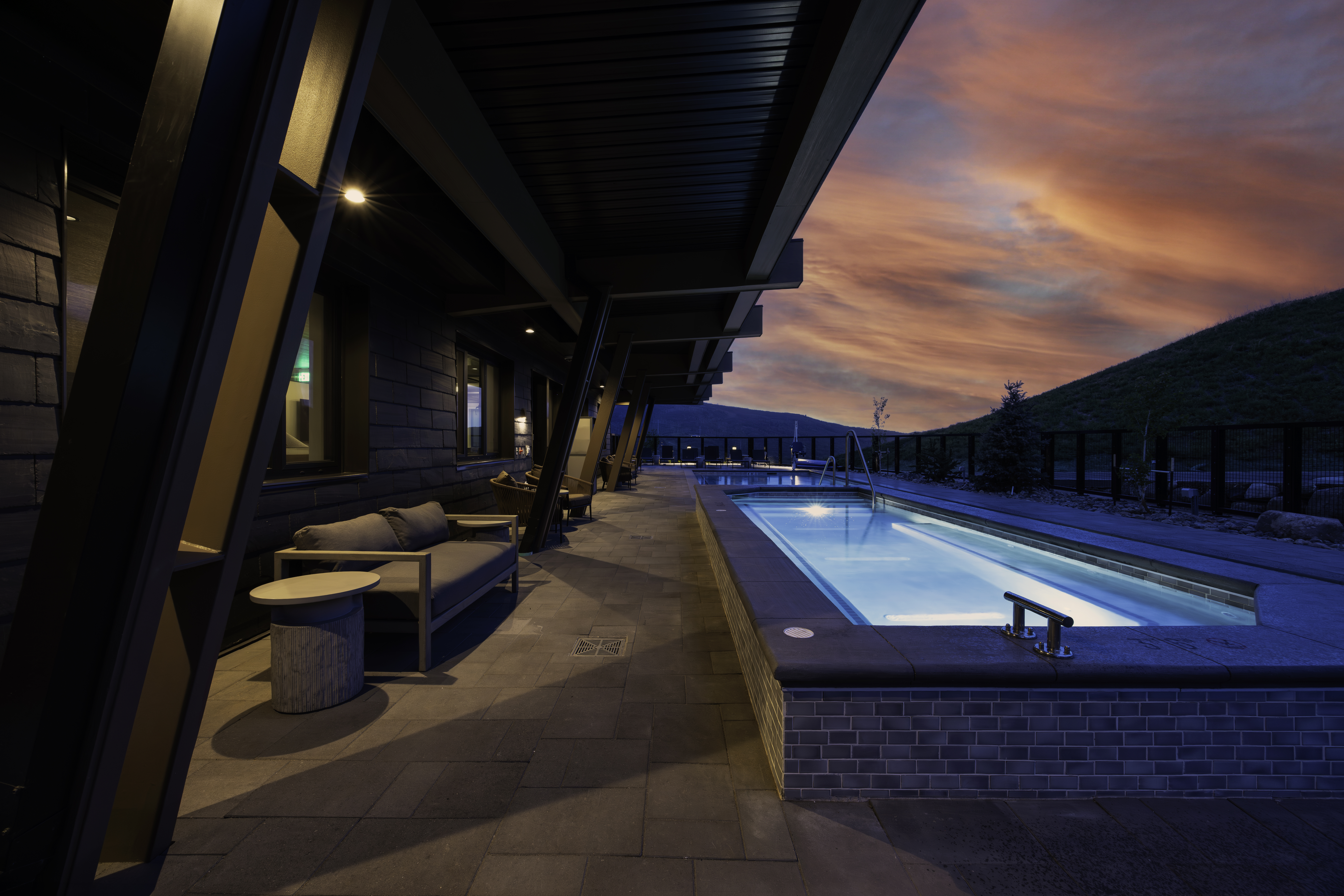 This striking outdoor patio features a sleek, rectangular lap pool illuminated with blue lighting, set against a dramatic sunset sky. The covered terrace is furnished with modern, minimalist seating and is framed by bold, angled structural supports that create a sophisticated, contemporary aesthetic. The scene captures a serene, high-end atmosphere perfect for evening relaxation, with the pool area extending toward a distant, rolling landscape.