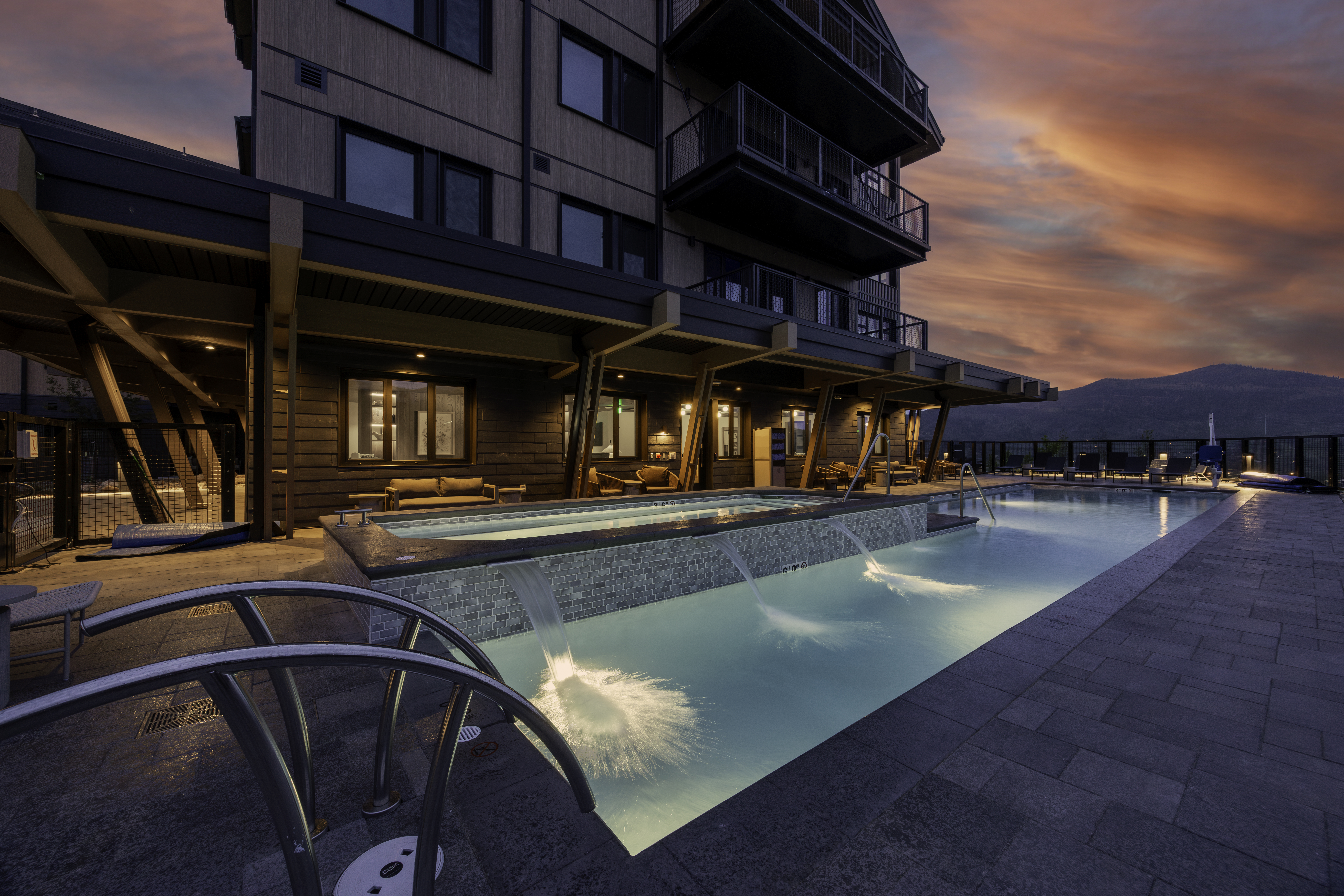 This cinematic shot captures a luxurious outdoor pool and spa area at dusk, set against a dramatic sunset sky. The scene features a multi-level pool design with cascading water features, illuminated by soft underwater lighting that creates a serene atmosphere. The surrounding patio is paved with stone, and the building's modern architecture is highlighted by warm exterior lighting, offering a sophisticated and inviting resort-style aesthetic.