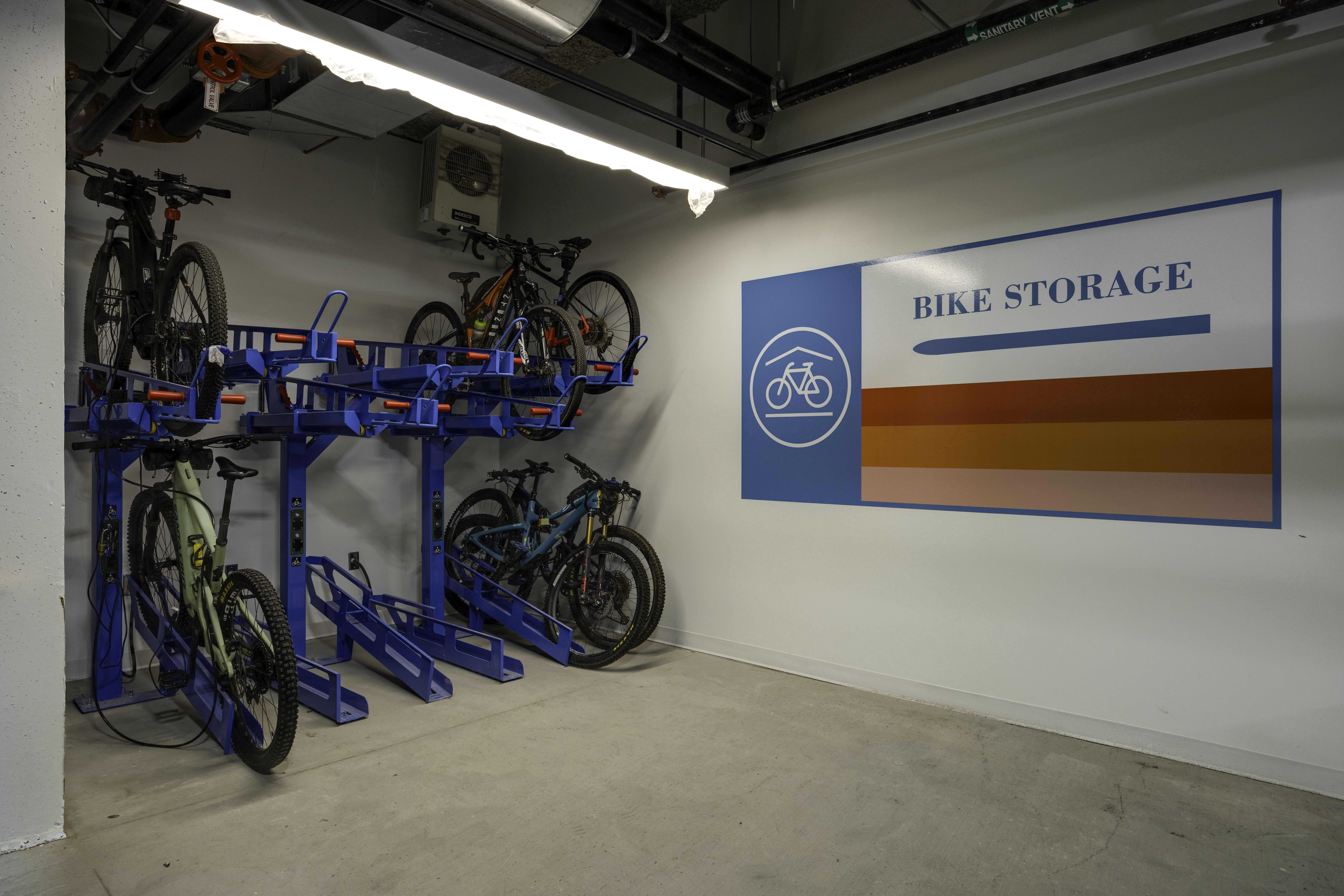 This image showcases a dedicated indoor bike storage area within a residential or commercial building. The space features a blue metal multi-level rack system holding several mountain bikes, set against a clean white wall adorned with a large, colorful 'BIKE STORAGE' graphic. The industrial-style ceiling with exposed pipes and lighting fixtures adds to the functional, utilitarian aesthetic of this convenient building amenity.