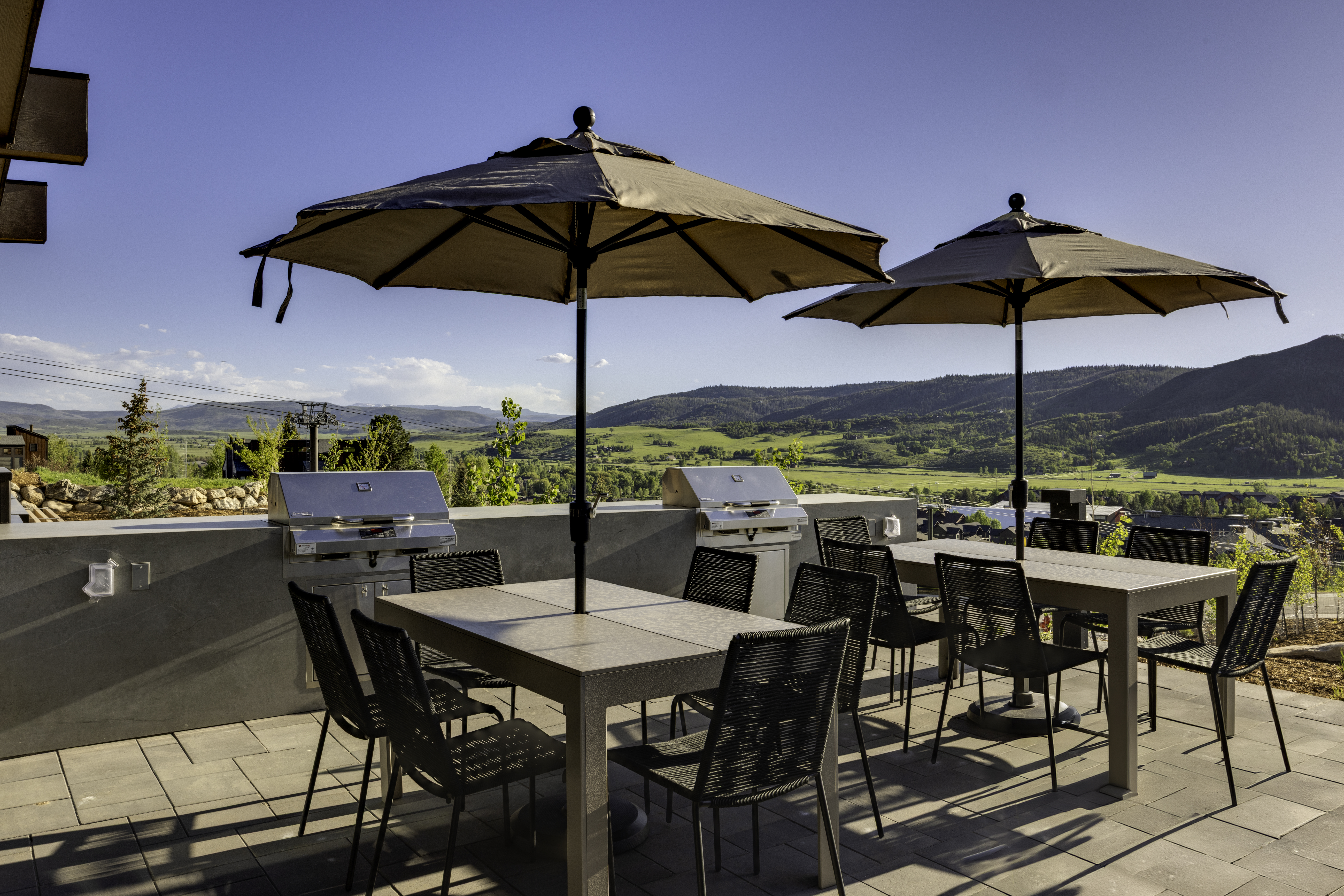 This elevated outdoor patio features two dining sets with matching umbrellas, providing a perfect space for al fresco dining with panoramic mountain views. The area is equipped with built-in stainless steel grills set into a concrete wall, emphasizing a functional and modern design. The paved surface and open layout create an inviting atmosphere for entertaining against a scenic, natural backdrop.