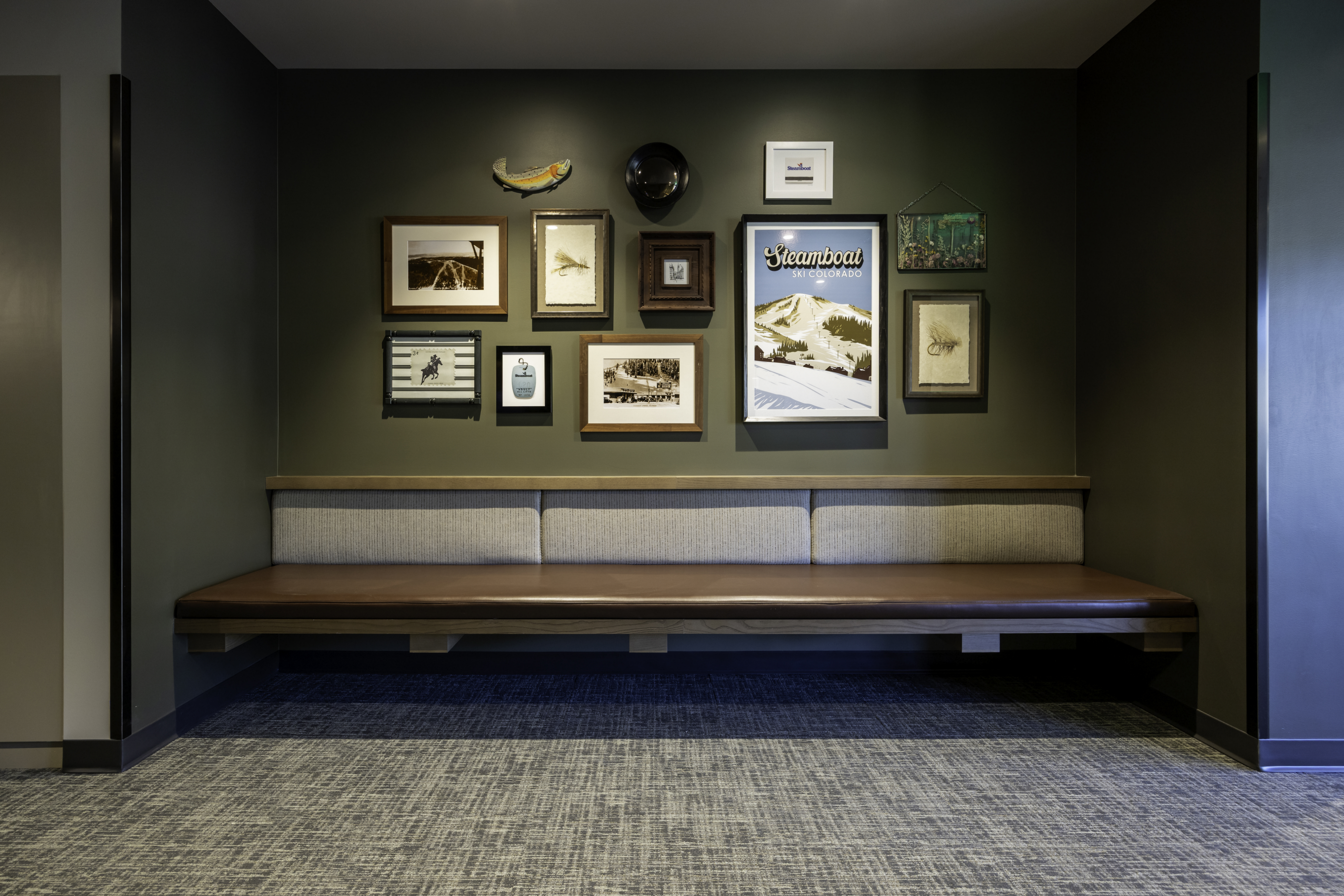 This interior space features a long, built-in bench with a dark leather seat and a light-toned fabric backrest, set against a deep olive-green wall. A gallery wall above the bench displays a curated collection of framed art, a decorative fish sculpture, and a circular object, creating a sophisticated and inviting focal point. The scene is captured from a straight-on, eye-level perspective, emphasizing the clean lines and balanced composition of the seating area.